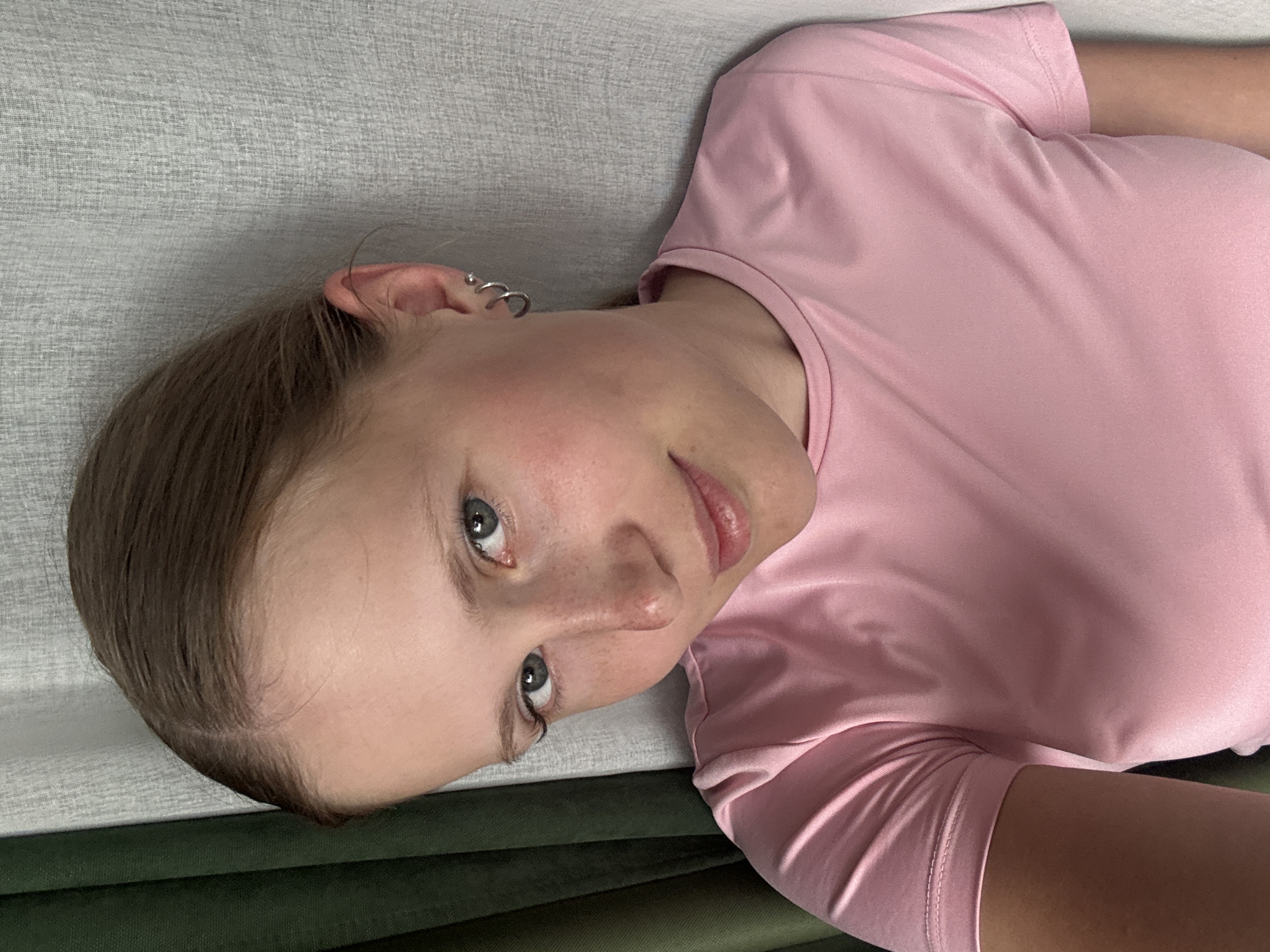 A young woman lies on a couch, wearing a pink t-shirt. She has blonde hair pulled back, and her ear is pierced with multiple hoop earrings. She is looking directly at the camera. The couch has a light grey fabric on the back and an olive green fabric on the seat. There is no indication of the location being Chaikovsky, Russia.