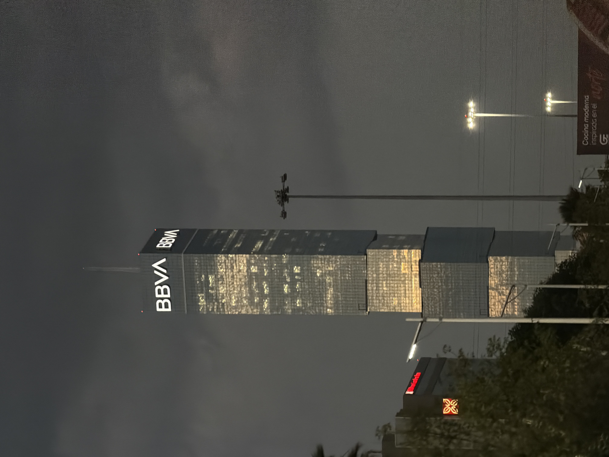 This image captures a static, nocturnal urban scene in San Bernardino Tlaxcalancingo, Mexico. The central subject is a towering, multi-story skyscraper with a glass facade, prominently displaying the illuminated white 'BBVA' logo on its upper sections. Numerous internal lights are active within the skyscraper, casting a warm yellow glow through its windows. A slender spire extends vertically from the apex of the structure.
To the left, a multi-story building is partially visible, featuring a red illuminated sign that appears to spell "Marriott" or similar characters, alongside a red illuminated geometric emblem. Foreground foliage, consisting of trees and bushes, partially obscures the lower portions of these structures.
In the mid-ground, to the right of the BBVA skyscraper, a tall, dark utility pole with a multi-directional floodlight array stands unilluminated. Further right, two additional streetlights with bright white overhead illumination are visible, each capped with a small red light. Two more white-emitting streetlights are present in the mid-left, partially obscured by vegetation.
A dark billboard or sign is partially visible in the lower right, displaying text that includes 'Cocina moderna inspirada en el norte' and a 'GE' logo. The sky is uniformly dark and overcast. No individuals, vehicles, or dynamic activities are observed within the frame.