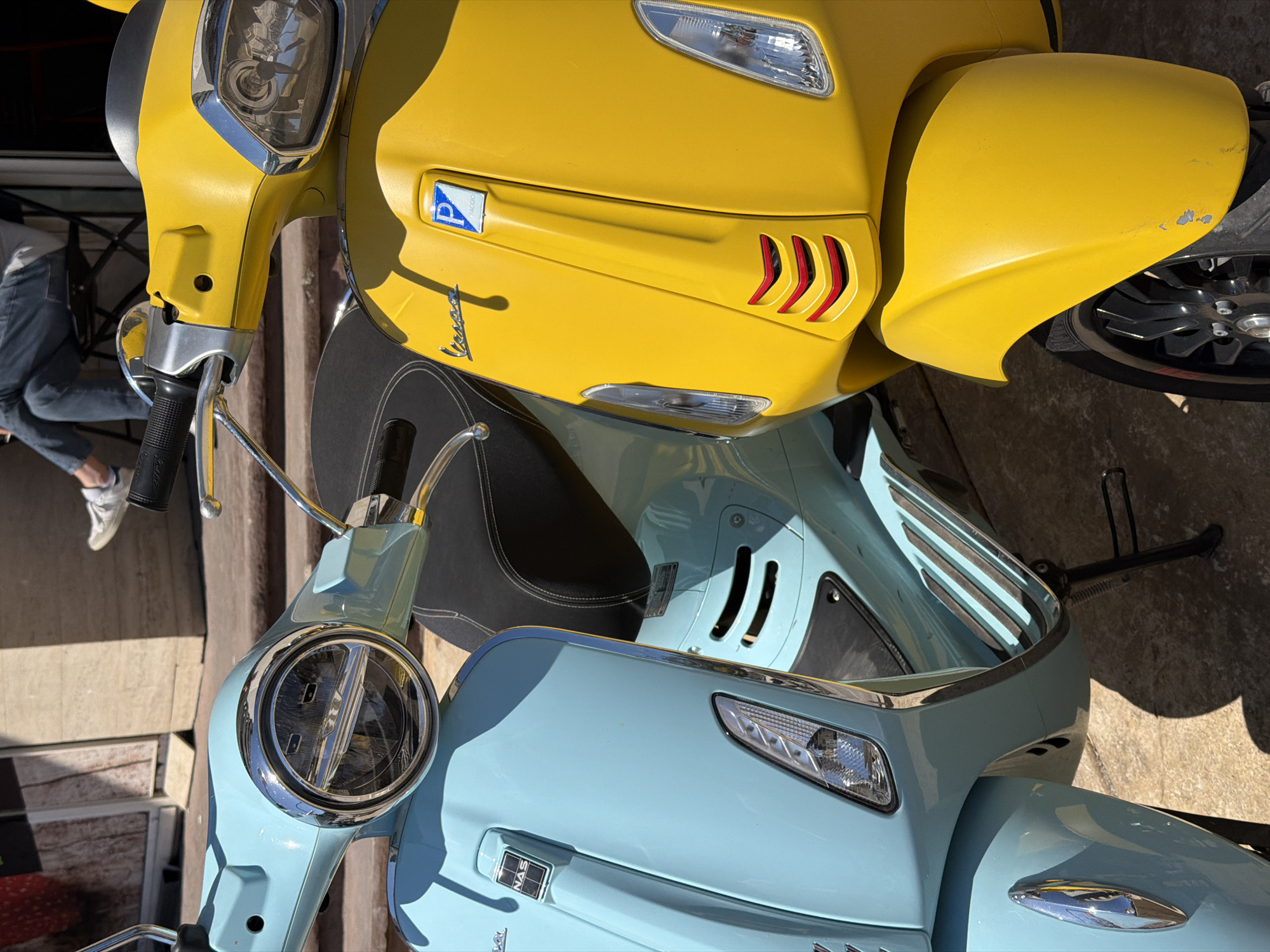 This image, taken in Budva, Montenegro, presents two parked scooters, a yellow one positioned above a light blue one, suggesting a vertical orientation of the photograph. Both scooters are classic Vespa models, with the distinctive "Vespa" script logo visible on their front panels.
The yellow scooter features a prominent chrome-bezeled circular headlight and clear turn signals. A blue Piaggio "P" emblem, with the word "PIAGGIO" underneath, is affixed to its front. Below this emblem, decorative red, slanted vent-like details are integrated into the front fairing. The yellow front fender shows noticeable signs of wear, with scuffs and paint chips revealing the underlying gray material. The scooter has a black seat with white stitching. Partially visible on the left, a person's lower legs and feet, clad in blue jeans and white sneakers, are in the background.
The light blue scooter, located below the yellow one, also has a chrome-trimmed round headlight and clear turn signal covers. A rectangular, metallic "NAS" emblem is visible on its front panel. Its seat is black, complementing the light blue body. This scooter is resting on a black kickstand on the right side of the frame.
The scene is brightly lit by natural sunlight, casting strong shadows on the concrete or paved ground, indicating clear weather during the daytime. The overall impression is of two colorful, classic scooters parked outdoors.