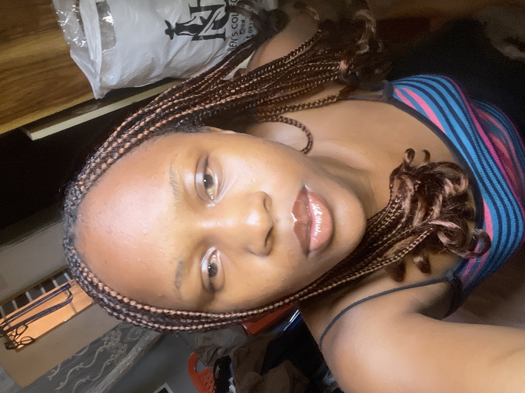 The image is a close-up selfie of a young Black woman with braided hair, taken indoors. She is wearing a tank top with horizontal stripes in blue, pink, and black. Her facial expression is neutral. In the background, there is a white plastic bag with black text and an image of a silhouette of a person wearing a hat. To the left of the woman, there are piles of clothing and a red object that appears to be a laundry basket. The lighting suggests an indoor setting, possibly a bedroom or living area. The overall impression is a casual, personal photograph.
This photograph was taken in Jos, Nigeria.