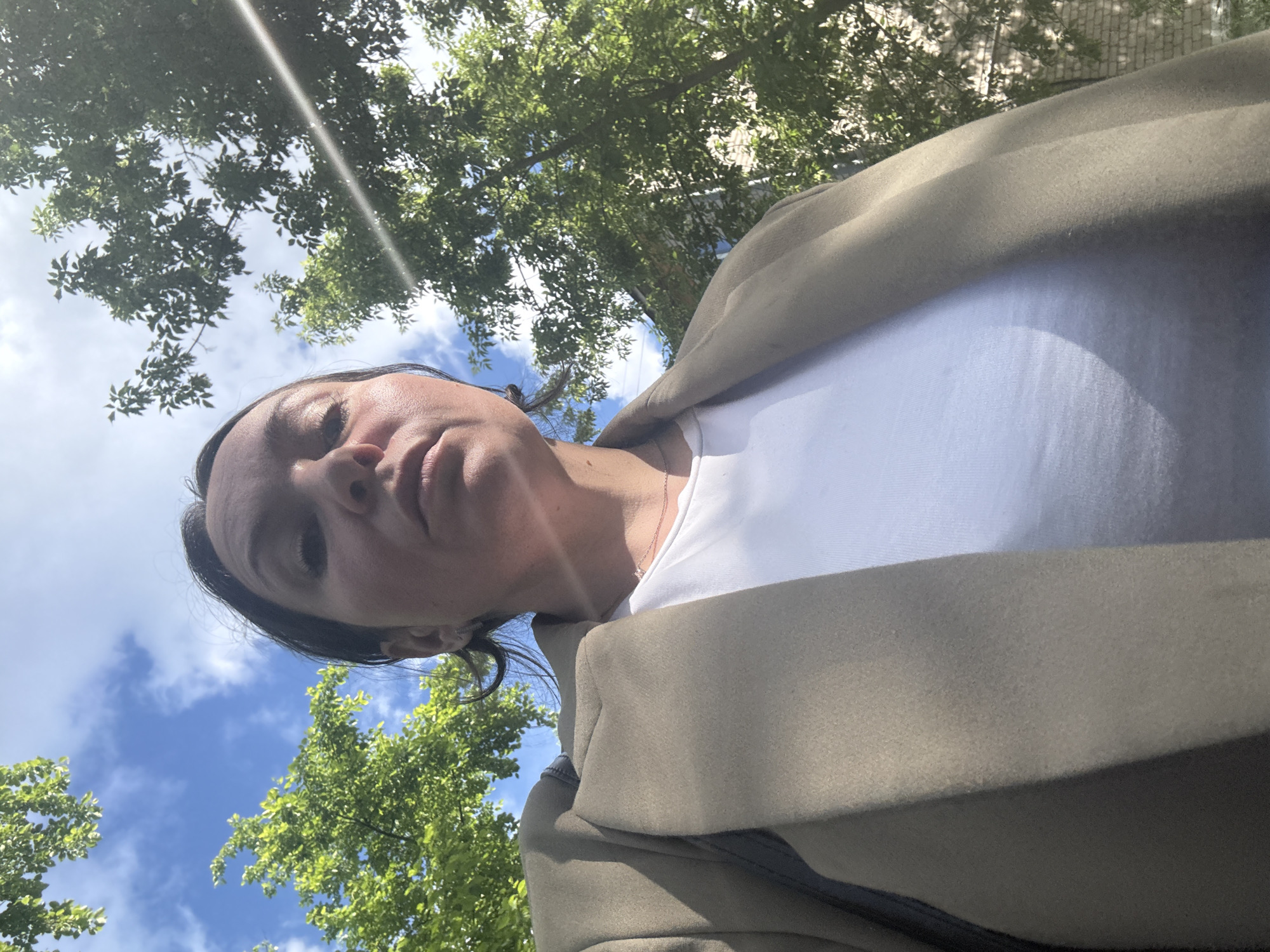 The image, correctly oriented vertically, presents a low-angle, close-up view of a person, likely a woman, outdoors. The shot is taken from below, looking upwards at the subject. Her face, from the chin to the top of her head, is prominent, with dark hair framing her features and a neutral to slightly serious expression. She is wearing a white t-shirt layered under a light brown or khaki jacket, and a delicate necklace is visible around her neck.
The background consists of a bright blue sky with scattered white clouds and vibrant green tree foliage. The strong sunlight indicates it is daytime, likely midday or afternoon, and casts some shadows on her face while also creating a noticeable lens flare in the upper portion of the image. The setting appears to be an outdoor natural environment, such as a park or a tree-lined area in Chaikovsky, Russia. No text is visible in the image.