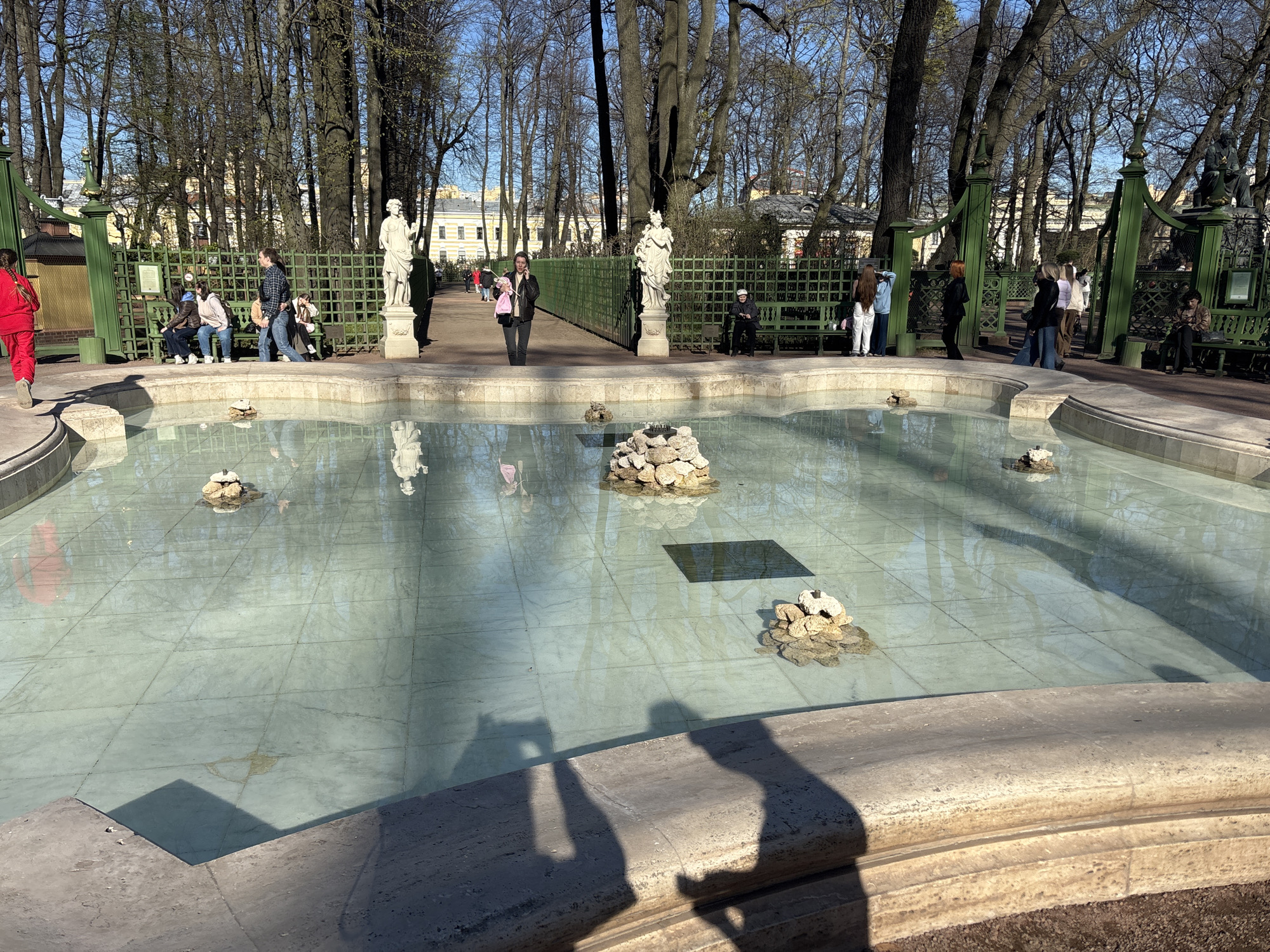 The image shows a rectangular, shallow pool or fountain with a light blue tiled bottom in the Summer Garden in Saint Petersburg, Russia. Several small piles of stones are placed in the water. The pool is surrounded by a low stone border. In the background, a green metal fence encloses the garden area with classical statues placed at intervals along it. People are seated on benches behind the fence, and others are walking along the path. Trees with bare branches surround the area. The sky is bright, indicating sunny weather. In the foreground, the shadows of two people holding a camera are cast upon the stone border of the pool.