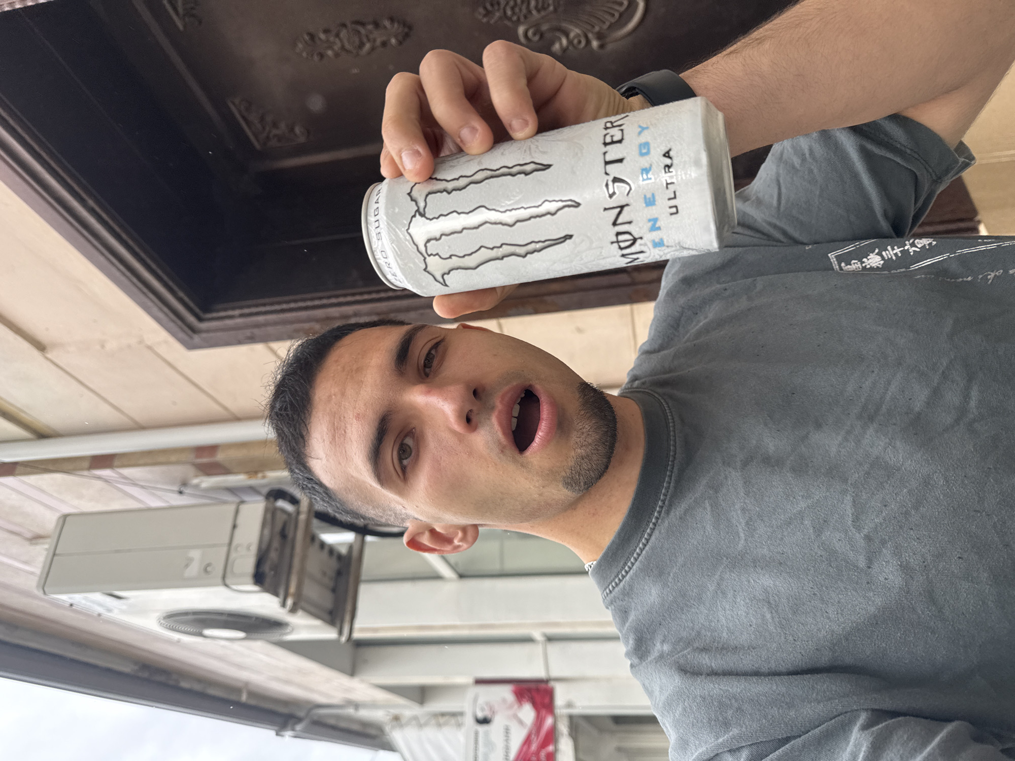 A man holds a can of "Monster Energy Ultra" drink in his right hand. He is wearing a gray t-shirt and a black smartwatch. The background shows elements of an urban environment, with a white air conditioning unit, a window or awning, and a dark brown architectural element that could be part of a building. The scene is most likely somewhere in Shumen, Bulgaria.