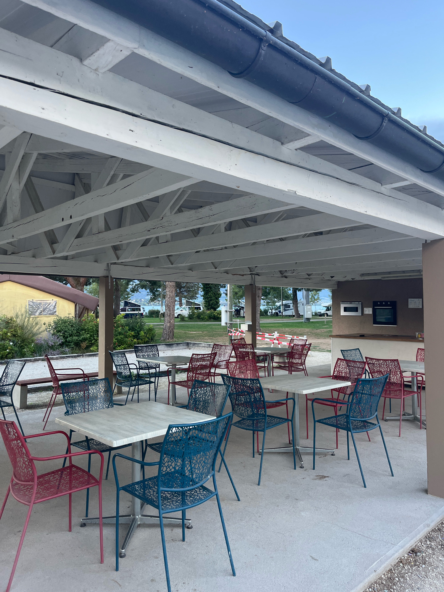 The image shows an outdoor covered seating area with multiple tables and chairs. The structure has a white painted wooden roof and support beams. The floor is concrete. In the foreground, there are several square tables with metal bases, surrounded by red and blue metal chairs with patterned seats and backs. In the background, more tables and chairs are visible, as well as a building with a yellow facade. Beyond the seating area, there is a grassy area with trees, a body of water, and mountains in the distance. Several white vans or recreational vehicles are parked in the background. The scene appears to be a common area in a campground or park. The location is Yverdon-les-Bains, Switzerland.