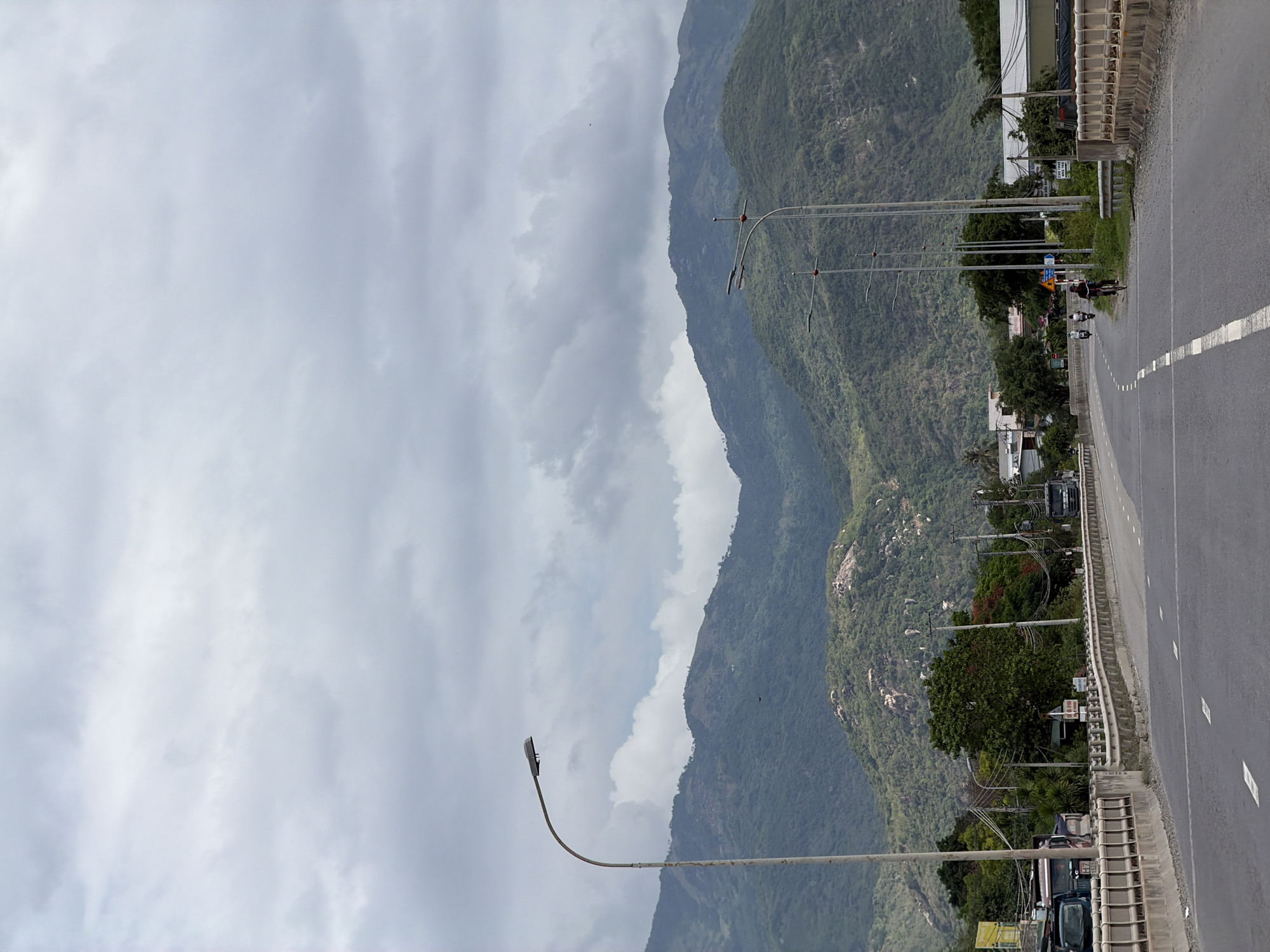 The image displays a panoramic view of a road winding alongside a lush, green mountain range under a heavily overcast sky. The scene is oriented horizontally, with the vast expanse of mountains dominating the left side and a paved road with an adjacent developed area on the right.
The sky is filled with dense, grey clouds, indicating an overcast daytime. The mountains are rugged and covered in dense vegetation, appearing verdant and imposing. Some rocky formations are visible among the trees. Towards the foreground and right side of the image, an asphalt road with clear white lane markings stretches into the distance. A white dashed line separates what appear to be two lanes, while a solid white line marks the shoulder.
A sturdy concrete guardrail runs alongside the road, separating it from the lower terrain and scattered buildings. Several streetlights with long, curved arms extend over the road, accompanied by multiple power lines supported by utility poles. Along the roadside, there are a few buildings, some partially hidden by trees, suggesting a degree of human settlement or commercial activity. A couple of motorbikes or scooters with riders are visible on the road, indicating light traffic. One motorbike is more prominent in the mid-ground.
The overall impression is one of a modern road infrastructure carved through a natural, mountainous landscape, typical of regions like Cam Ranh Ward, Vietnam, where natural beauty coexists with development. No legible text is visible on any signs.