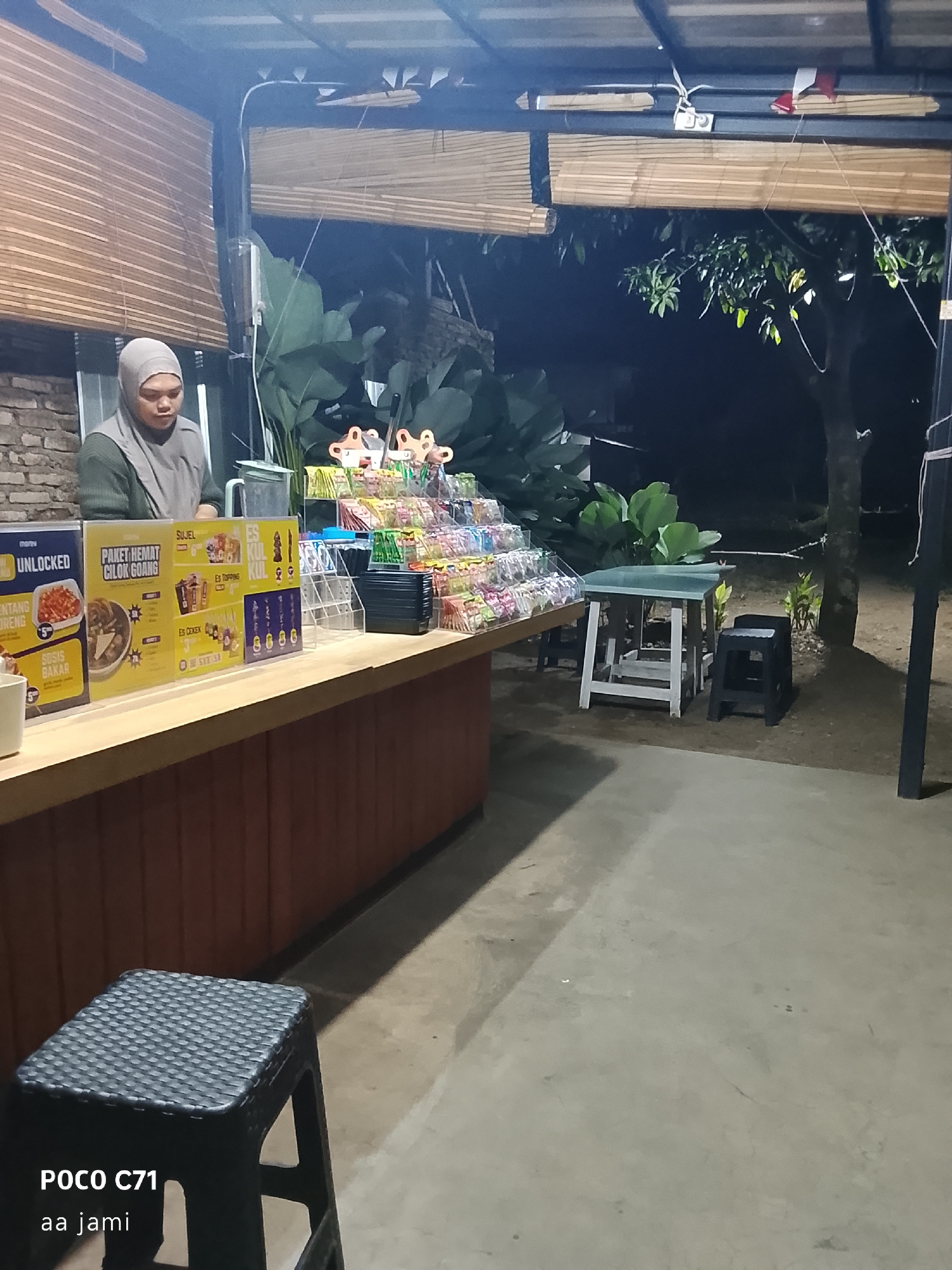 The image depicts an outdoor food stall at night, likely in Tasikmalaya, Indonesia. A person wearing a hijab stands behind a counter filled with various snacks and drinks. In front of the counter are signs advertising menu items such as "Paket Hemat Cilok Goang" and "Sosis Bakar." A small table with two stools is set up to the right of the stall. The background shows lush greenery and trees, suggesting a natural or semi-rural setting. The overall atmosphere is casual and informal, consistent with a street food vendor. The floor is concrete, and a black plastic stool is visible in the foreground. The lighting suggests it is nighttime.
