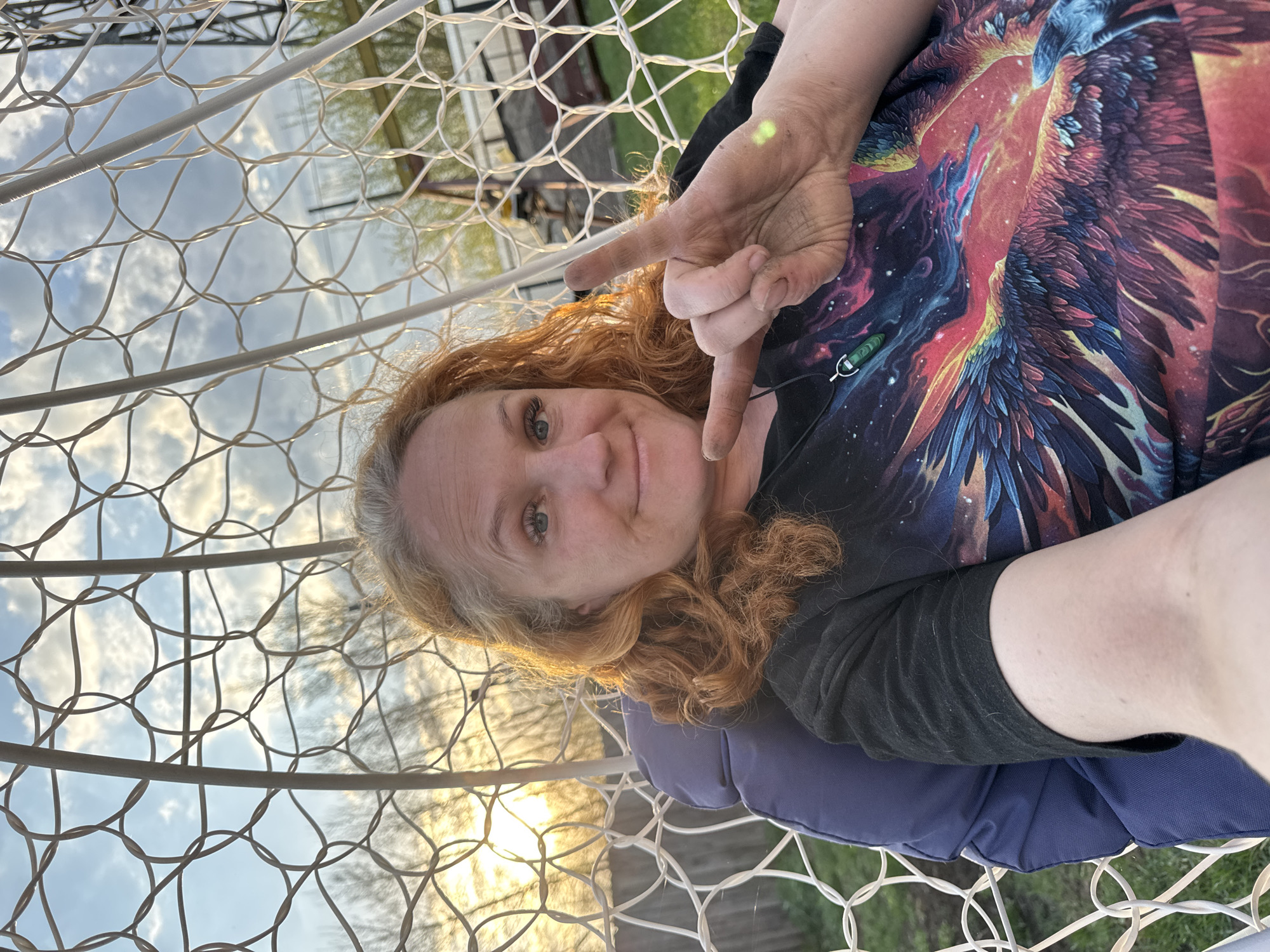 A woman with reddish-blonde hair is lying in a white wire hanging chair outside, possibly at sunset, based on the lighting. She is making a hand gesture that appears to be the letter "L", wearing a black shirt with a bird/galaxy design and a black necklace with a green stone. A blue cushion is behind her. The background shows trees and a partially visible building through the wire frame of the chair. The photo was taken in the city of Chaikovsky, Russia.