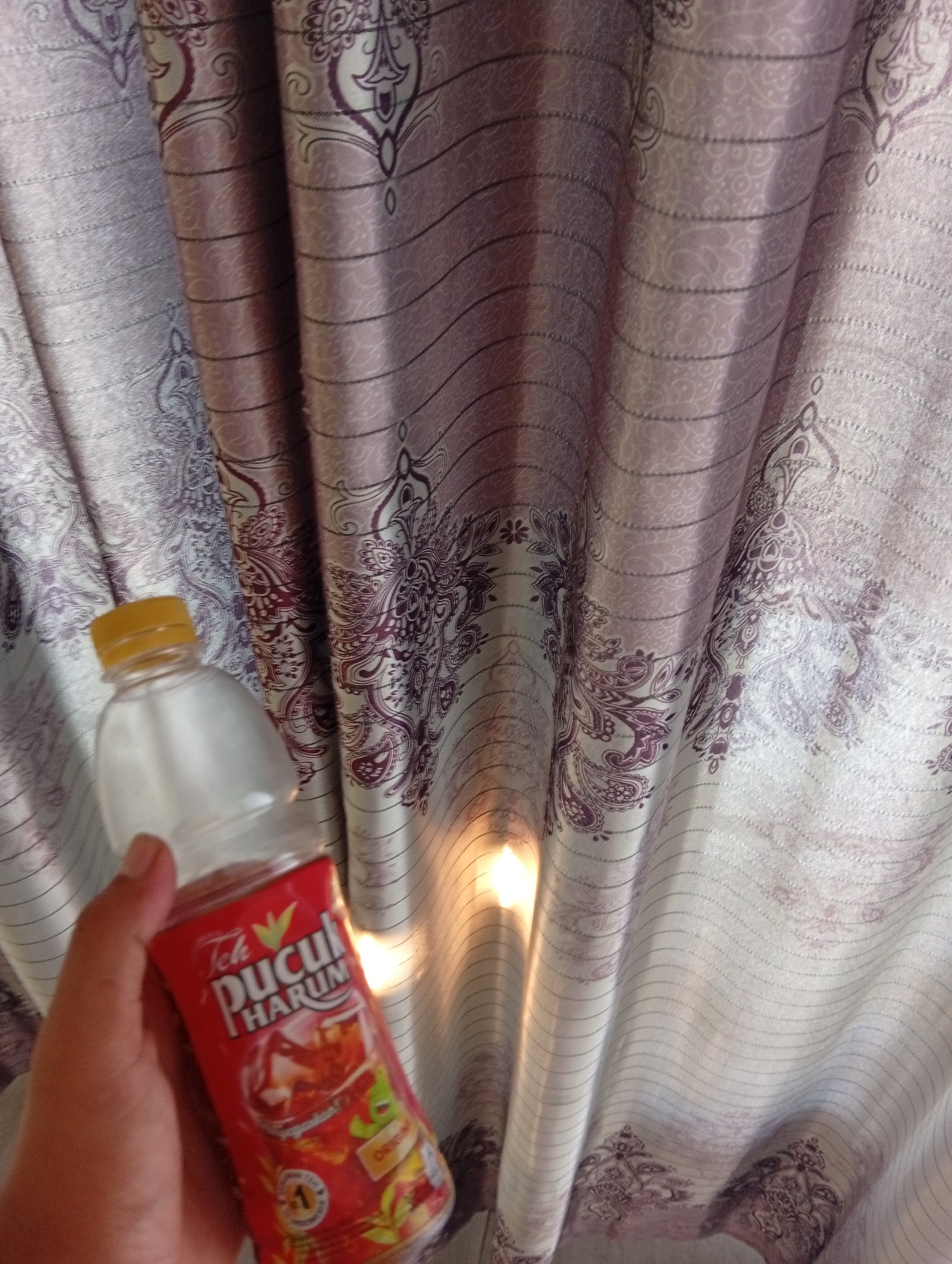 A hand holds a clear plastic bottle of Teh Pucuk Harum iced tea in front of a purple and white patterned curtain. The bottle is partially filled with a dark liquid and has a yellow cap. The curtain has a repeating paisley design and horizontal lines. Light reflects off the curtain, creating a bright spot. The scene appears to be indoors, likely in a home, given the presence of curtains and the casual way the bottle is held. The location is not specified in the image.