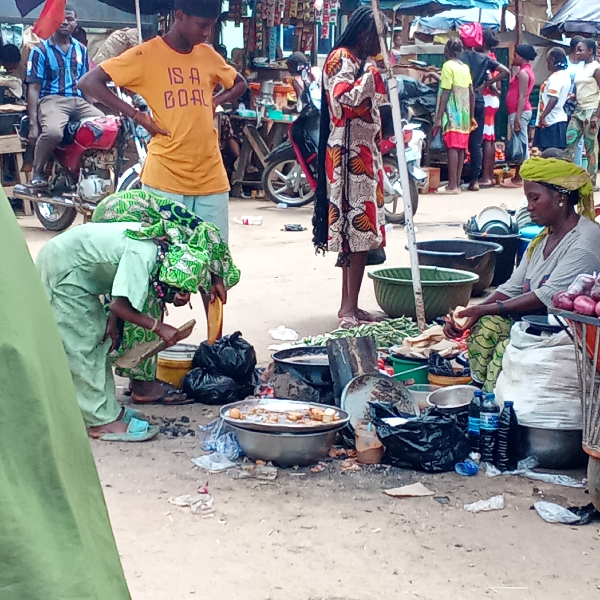 A bustling market scene in Akwanga, Nigeria. A woman in a light green outfit bends over, attending to a cooking setup. A woman in patterned attire stands nearby. A man in an orange shirt with the words "IS A GOAL" stands with his hands on his hips. Another man in a blue striped shirt sits on a red motorcycle. A woman with a yellow headscarf sits at a market stall, handling food. Other people are seen milling in the background. There are various items for sale in the foreground including food in a metal bowl, bottles of oil and water. Debris litters the ground.