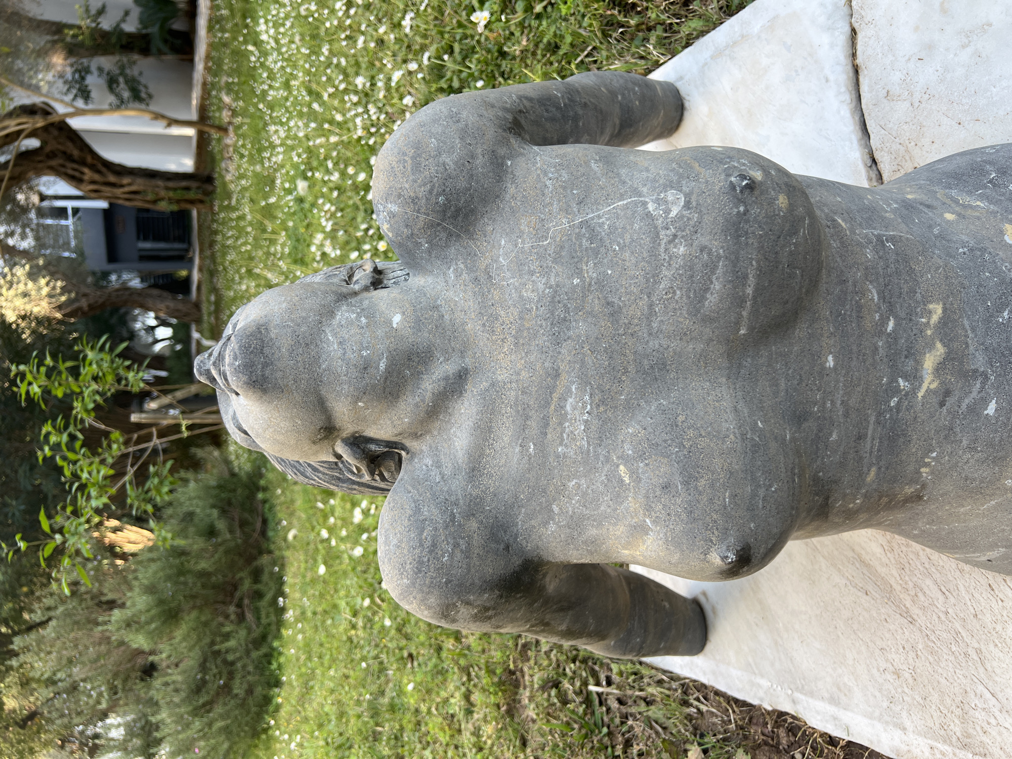 The image depicts a weathered, gray stone statue of a nude woman, likely an art installation or sculpture, set outdoors. The statue is positioned on a white, possibly marble, platform or base. The background includes a lawn with small white flowers, shrubbery, and a building in the distance with a tree. The statue is located in the city of Petrovac na Moru, Montenegro.