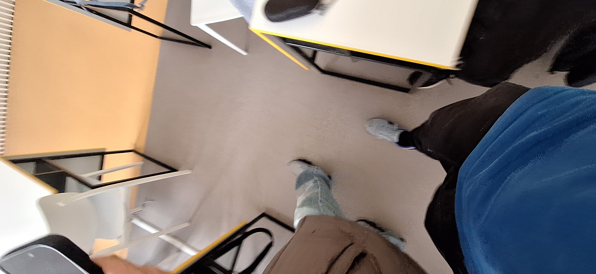 The image shows an indoor scene in what appears to be a classroom or office setting. There are desks and chairs visible. The floor is a neutral light gray. A person is standing in the lower right corner of the frame, wearing dark pants and a bright blue shirt. The image may have been taken by another person, as a hand and phone or device can be seen in the lower left corner.
The scene may be in Burgas, Bulgaria, but the image does not provide enough information to verify the location.