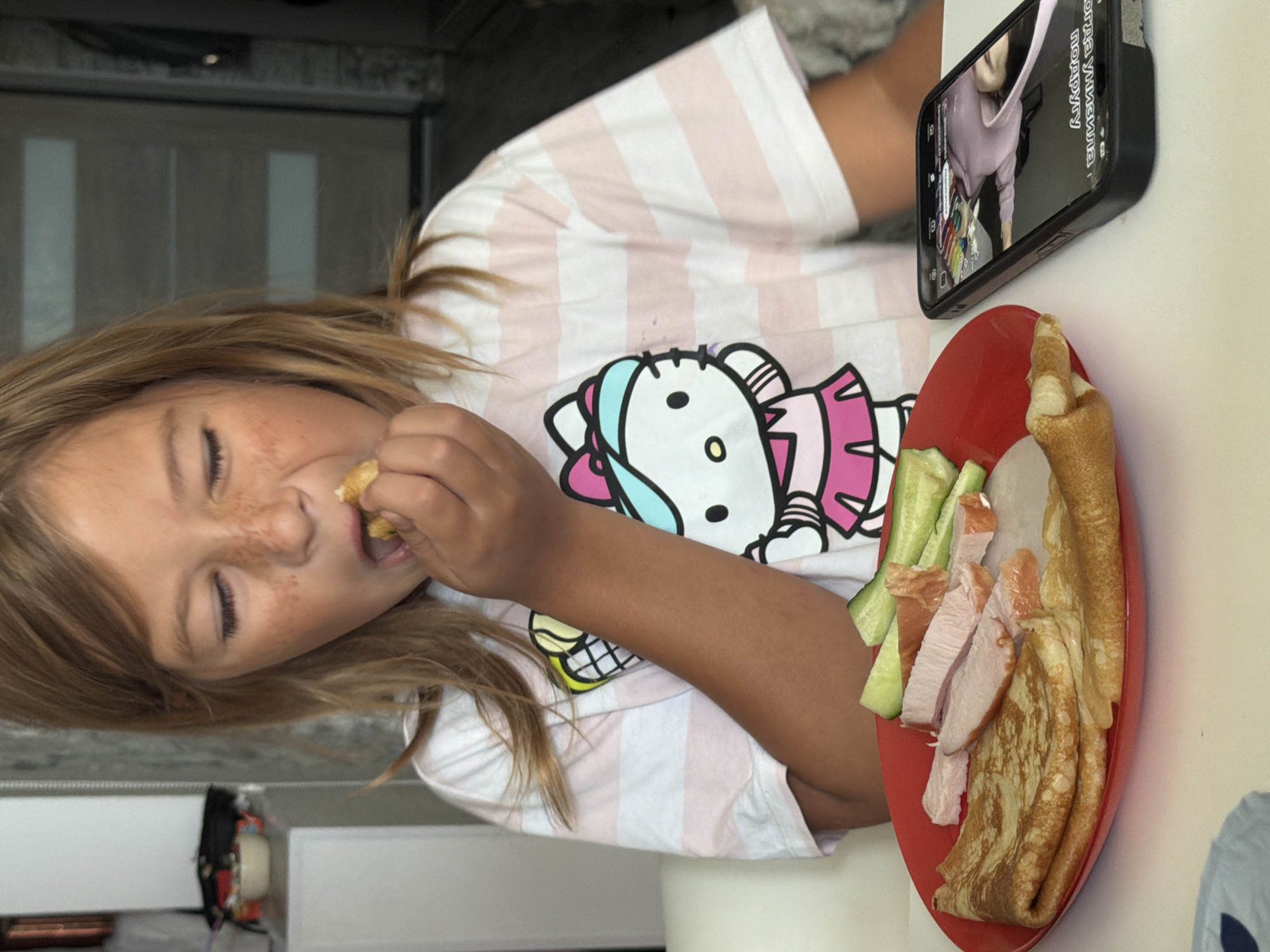 A young girl with blonde hair and freckles is eating. She is wearing a pink and white striped t-shirt with a Hello Kitty graphic. In front of her is a red plate with pancakes, sliced ham, and cucumber. A smartphone is lying on the table next to the plate, displaying a video of a person. The setting appears to be indoors, possibly a kitchen or dining area. The city of Sevastopol, Ukraine, is a port city on the Crimean Peninsula.
