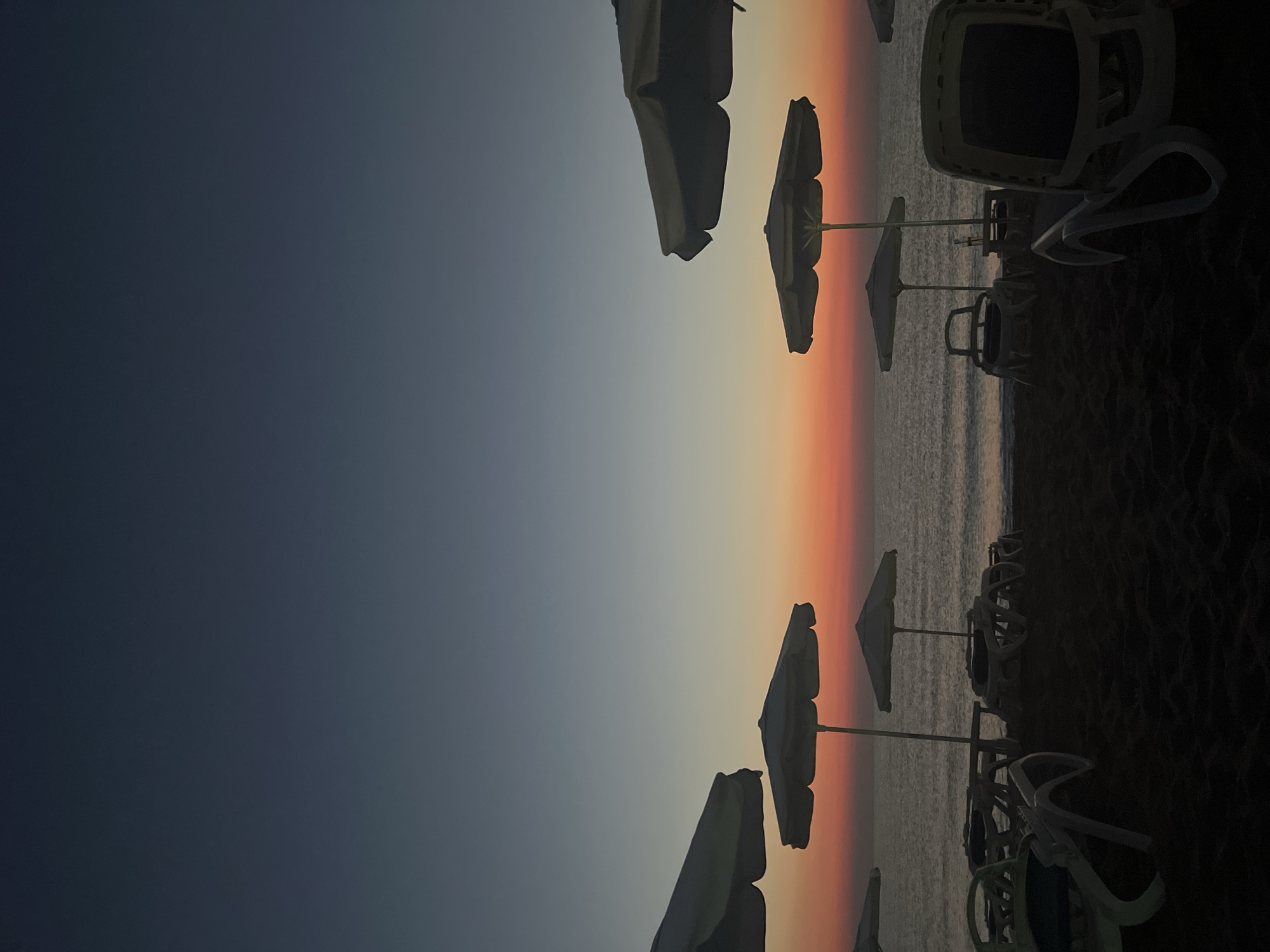 This image depicts a twilight scene on a beach in Lozenets, Bulgaria. In the foreground, several empty lounge chairs are scattered across the dark sand, some with white plastic frames. Behind them, a row of white beach umbrellas stand open, casting shadows. The sea stretches out behind the beach, its surface rippling gently. The sky above is a gradient of dark blue at the top, transitioning through shades of pale blue and orange to a vibrant red near the horizon, indicative of a sunset or sunrise. The overall atmosphere is serene and peaceful, with the beach appearing deserted.