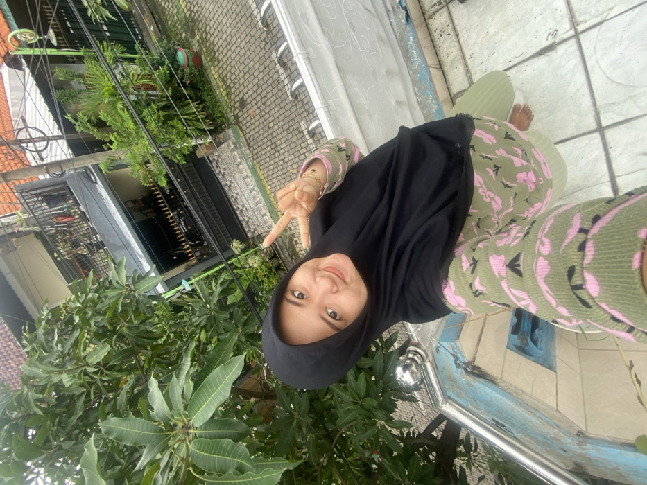 A woman wearing a black hijab and a green and pink patterned outfit stands on a tiled surface. She flashes a peace sign, smiling towards the camera. Lush green foliage is on the left, with a glimpse of buildings and overhead wires in the background. A railing runs along the bottom, and more tiled surface stretches out behind her.