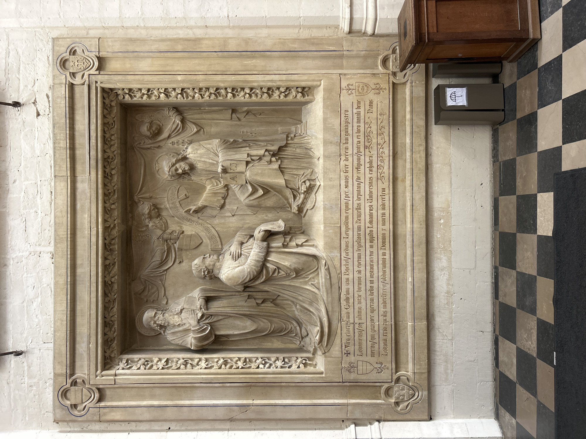The image shows a detailed stone relief depicting four figures and Latin inscriptions, located on a wall in Leuven, Belgium. The relief is framed by decorative elements and set against a plain stone background.
At the center of the relief, a seated figure, presumably Christ, with a halo, holds an open book bearing the letters "AW". To his left, another seated figure, likely a scholar or dignitary, is gesturing towards Christ with his right hand. To the left of the seated figures stands a third figure, a bearded man with a halo, holding his hands together. To the right of Christ, a fourth figure, cloaked and with a halo, stands behind him. The figures are rendered in a classical style with flowing drapery.
Below the relief are two lines of Latin text. The first line reads: "Viro clarissimo Guillelmo van Borkel ordmis Leopoldini Lovanienst," and the second line continues with more text that is partially obscured and difficult to read. There are also crests and decorative elements flanking the text.
The relief is situated on the interior wall of a building, with a checkered black and white tiled floor visible in the foreground. To the right of the relief, there is a brown wooden donation box or cabinet, and a modern grey trash receptacle with an umbrella symbol on a white label. The wall behind the relief is constructed of white brick. The overall impression is of a historical or religious monument within an institutional setting.