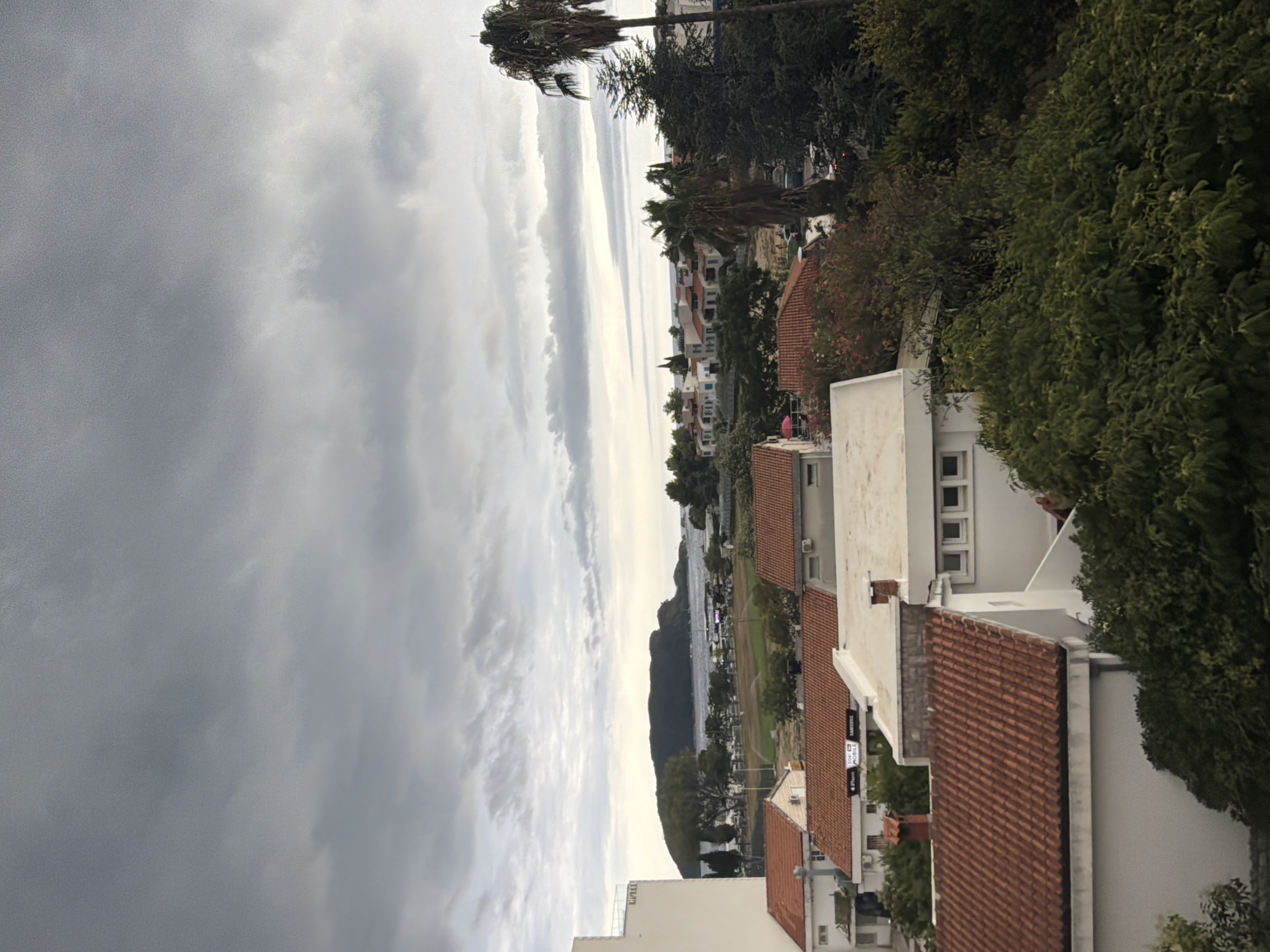 The image depicts a cloudy sky over a coastal town. In the foreground, there are several white buildings with terracotta tiled roofs. To the left, a tall, modern white building, possibly a hotel, is partially visible. Green trees and foliage surround the buildings. In the middle ground, there is a landscape with more white buildings, some with blue accents, nestled amongst trees. Beyond that, a body of water, likely the Adriatic Sea, is visible, with a large, dark, mountainous island or peninsula in the distance. The sky is filled with a variety of grey clouds, suggesting overcast or stormy weather. The scene is a view from an elevated position, looking towards the sea and the town of Budva, Montenegro. There are no people or clear actions discernible in the image.