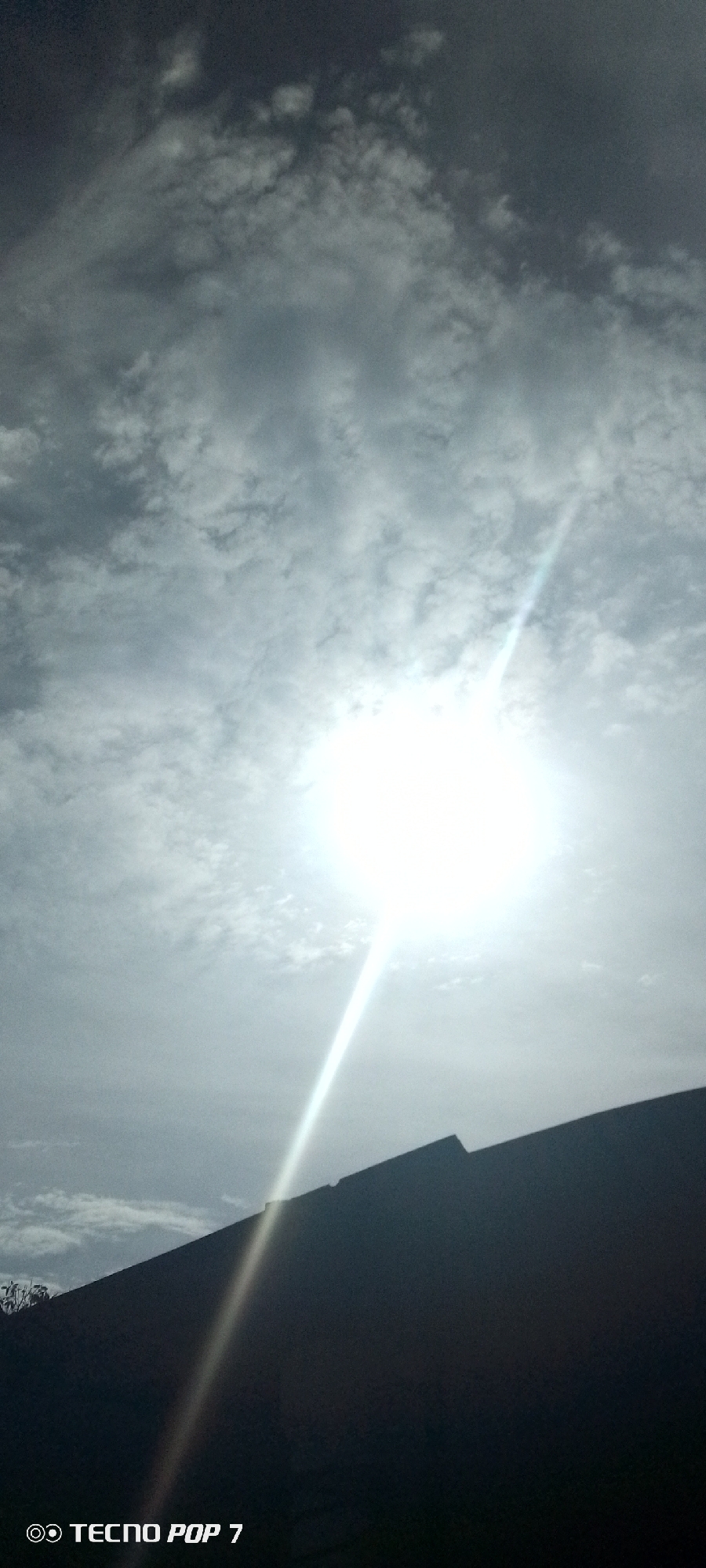 The image shows a bright sun obscured by thin, textured clouds in Altagracia, Venezuela. A dark, angular structure, likely a building roof or wall, occupies the lower portion of the frame. A bright ray of light emanates from the sun, piercing through the clouds and reaching towards the bottom. The "TECNO POP 7" watermark is visible at the bottom left corner.