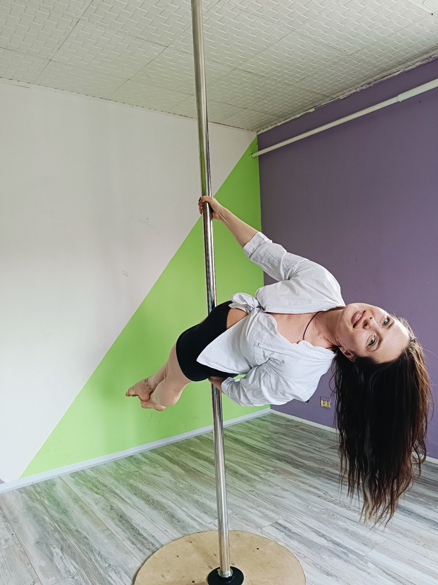 A woman is performing pole dance. She is wearing a white shirt and black shorts. Her hair is long and brown. The background is a room with white and green walls on the left, and purple walls on the right. The ceiling is textured and white. A metal pole stands on a round wooden base. The location is a room in Rusalevka, Russia.