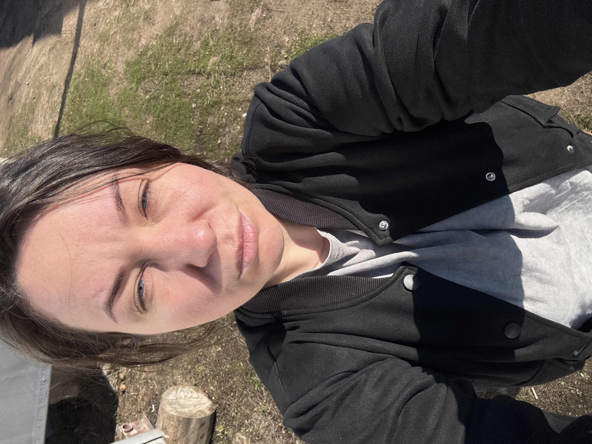 The media file is a selfie photograph taken outdoors of an individual in Chaikovsky, Russia. The image is rotated, showing the subject from a high angle, looking down towards the camera.
The central subject is a person with dark hair, fair skin, and partially closed blue or grey eyes, possibly due to bright sunlight. They have a neutral to relaxed expression. A small, delicate chain is visible around their neck. They are dressed in a black jacket, likely a varsity or bomber style, featuring a ribbed collar and cuffs, and snap buttons along the front. Underneath the jacket, a light grey t-shirt is visible.
The background depicts an outdoor setting characterized by a mixture of dry, light brown earth and patches of green grass. A distinct, long shadow is cast across the ground from an unseen object to the left, indicating bright sunshine. To the lower right of the frame, a cut log or tree stump is visible, along with some indistinct debris. The bright, direct light suggests it is daytime, likely late morning or early afternoon, with clear skies.
The activity taking place is a self-portrait (selfie), with the camera seemingly held above and slightly in front of the subject. There is no visible text in the image.