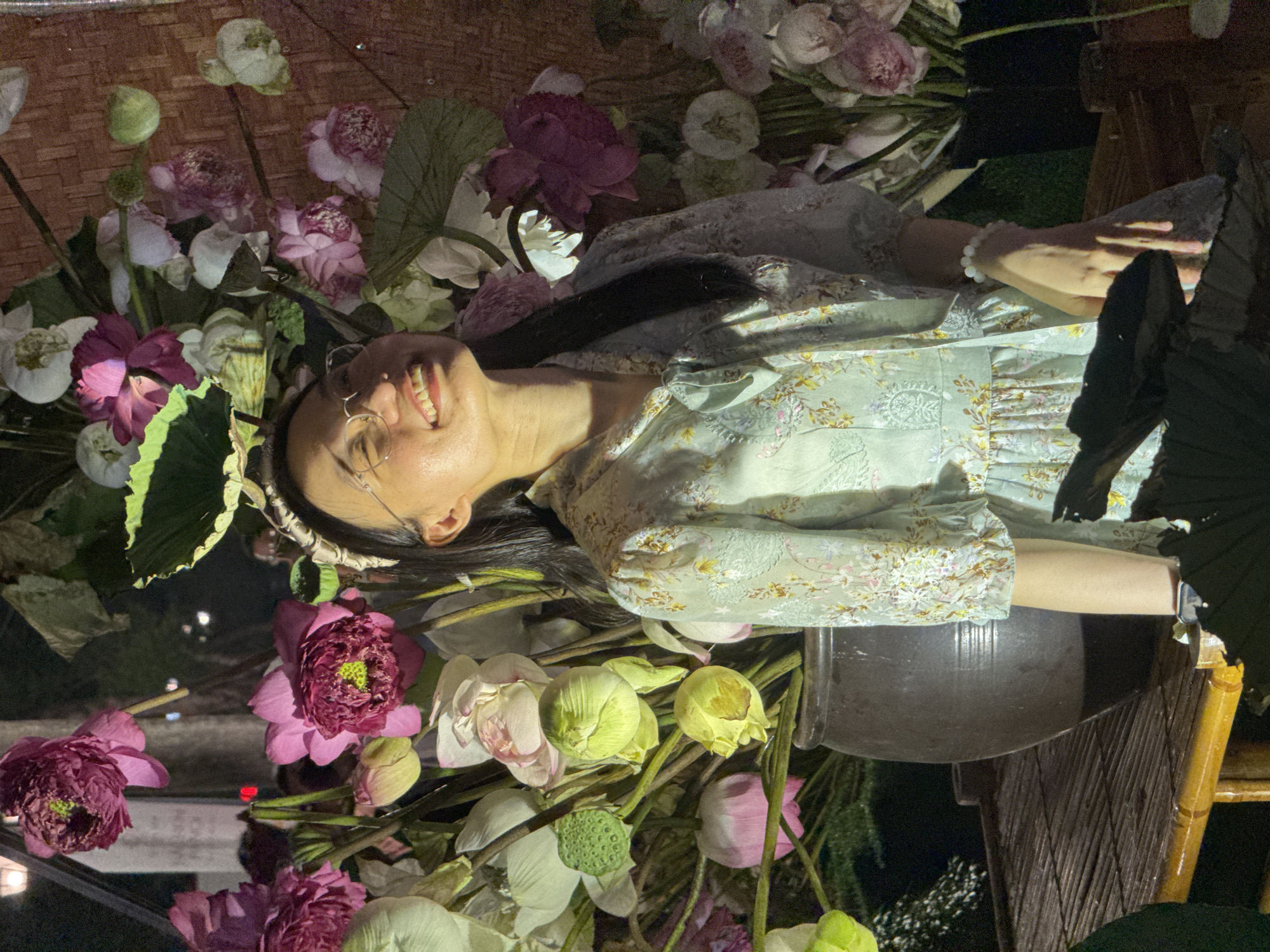 A young woman with glasses and a floral headband stands in front of a large arrangement of pink and white lotus flowers. She is wearing a light blue, floral-patterned dress and smiling up at something off-camera. The arrangement is in a dark vase on a wooden table. The scene appears to be at night, with artificial lighting illuminating the woman and flowers. The background shows a dimly lit urban environment, potentially a street in Hanoi, Vietnam, indicated by the presence of streetlights and the style of the woven bamboo backdrop.