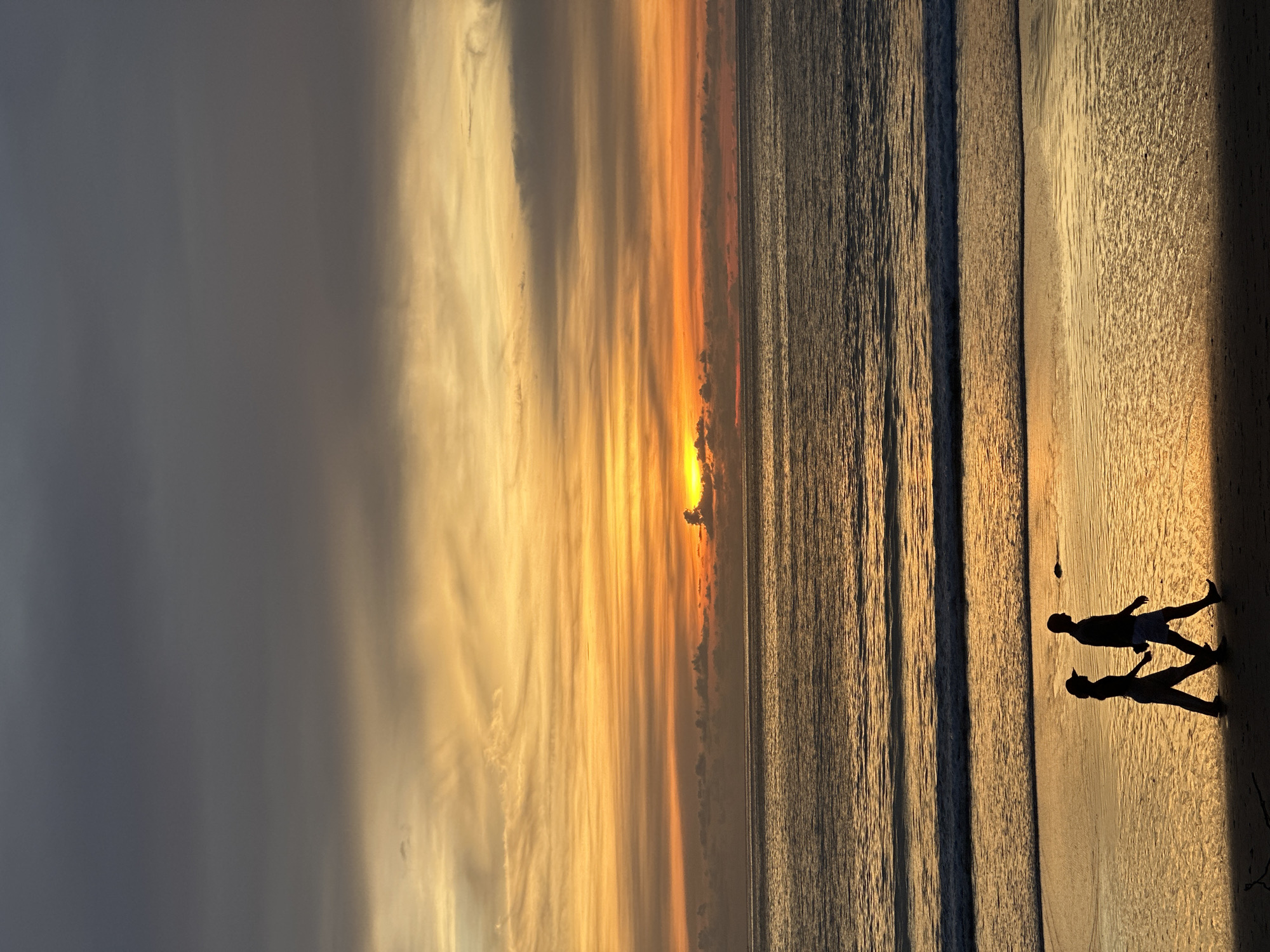 A vertical, full shot captures a silhouette of two people walking on a beach during a dramatic sunset over the ocean. The sky is filled with vibrant orange and yellow hues from the setting sun, contrasted by darker grey clouds at the top. The ocean reflects the colors of the sky, with gentle waves reaching the shore. The two figures, a man and a woman, are walking side-by-side along the wet sand, their bodies in profile. The man is taller and wears shorts, while the woman is shorter and appears to be wearing pants and a hat. The overall scene evokes a sense of tranquility and natural beauty. The location is described as Unknown, Unknown.