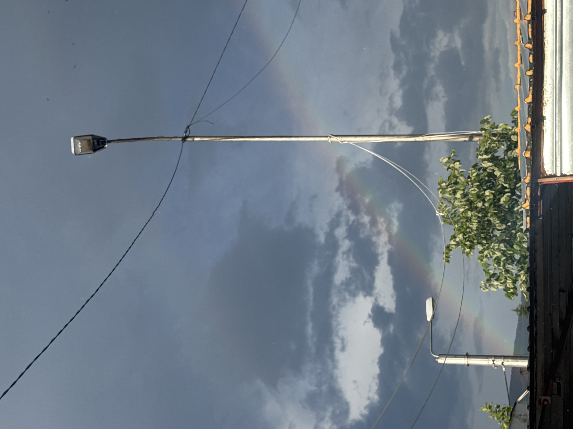 The image shows a cloudy sky with a faint rainbow visible. A street light pole with a light fixture is in the foreground. Electrical wires stretch across the sky. Part of a building with a corrugated metal roof and some foliage are on the right side of the image. It is unknown whether or not the city of Sliven, Bulgaria is depicted in the image.