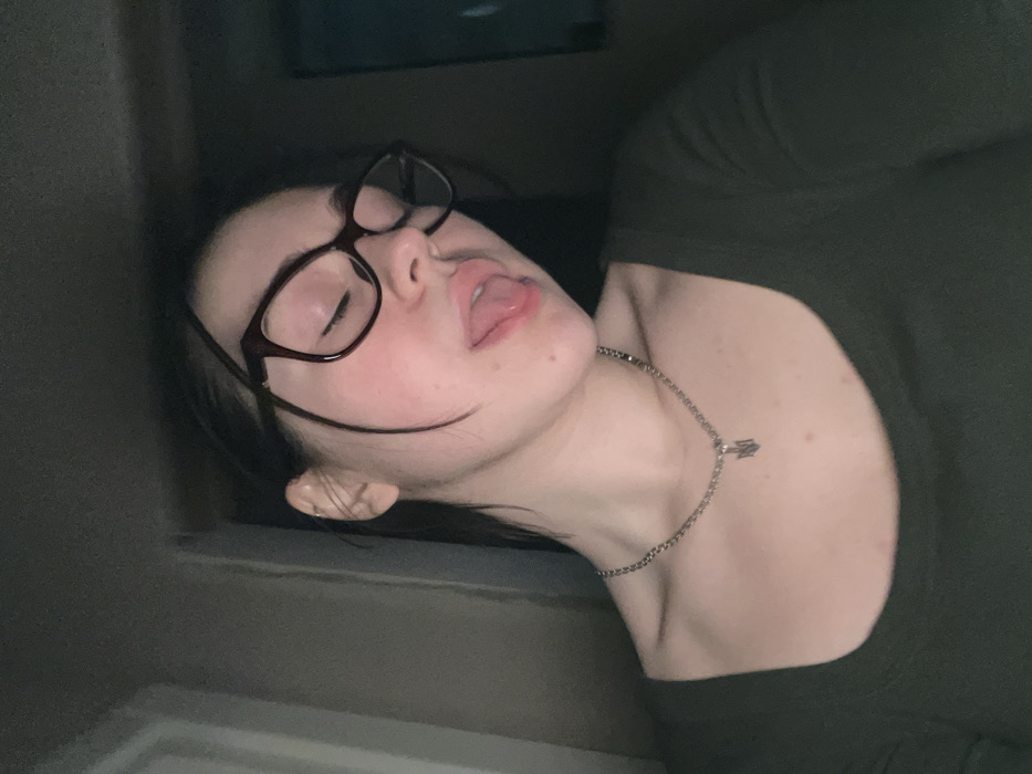 The image displays a female individual from the chest up in a dimly lit indoor setting. She has her eyes closed and her tongue slightly extended from her mouth. She is wearing dark-framed eyeglasses and a dark green, long-sleeved top with a scoop neckline. A silver-colored chain necklace with a trident-shaped pendant is visible around her neck. A small, gold-colored hoop earring is present on her left ear. The background is dark and indistinct, offering no specific details about the room. There are no visual elements within the image to identify the location as Rafailovići, Montenegro.