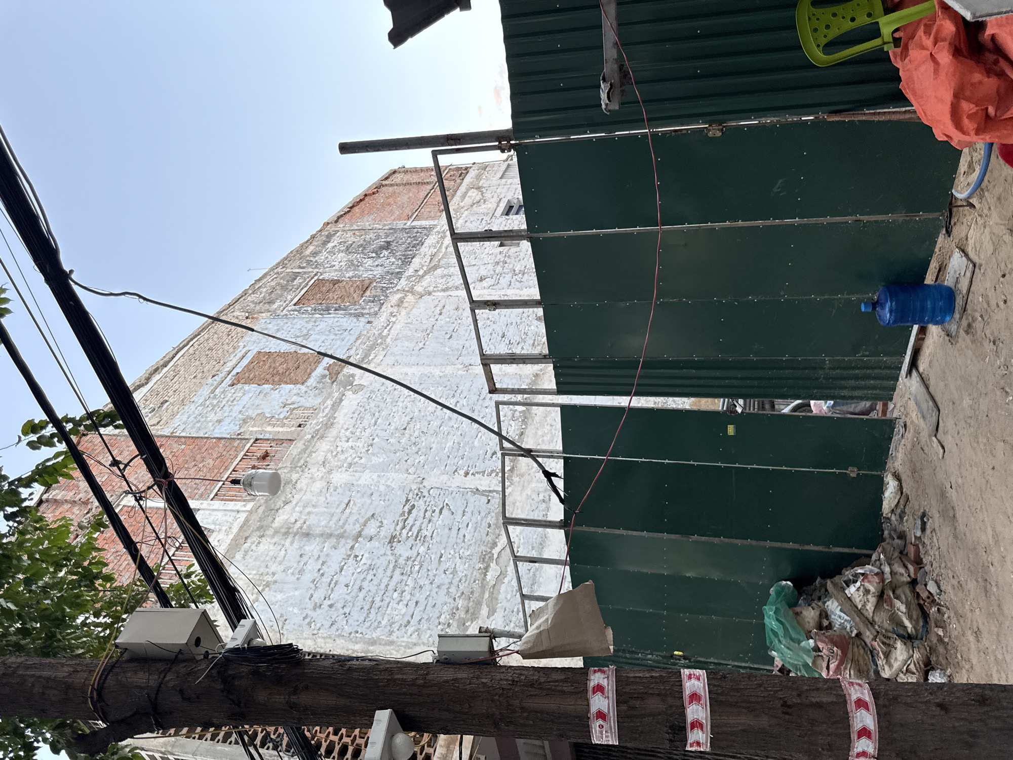 This image depicts a construction site in Nha Trang, Vietnam. A partially constructed or dilapidated building with exposed brickwork and peeling white paint dominates the background. The building has several boarded-up windows. In the foreground, a green corrugated metal fence obstructs the view of the site. Piles of debris, including sacks of material and loose bricks, are scattered on the sandy ground near the fence. Electrical wires hang precariously from a utility pole on the left side of the image, which is wrapped with red and white caution tape. A large blue water jug sits on a wooden plank in front of the fence, and a bright green plastic chair is visible on the right. The sky above is clear and blue, suggesting daytime.