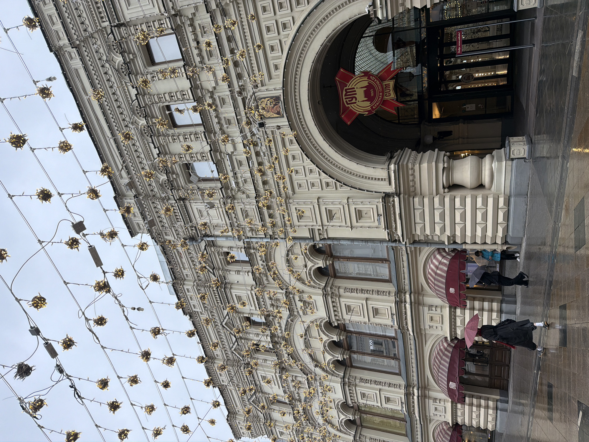 The image depicts a view of the GUM department store in Moscow, Russia, partially rotated. The architecture is visible, adorned with decorative lighting. The storefronts have awnings, and at least two people with umbrellas are walking by. The ground appears wet, suggesting rain. A red emblem hangs above the central entrance.