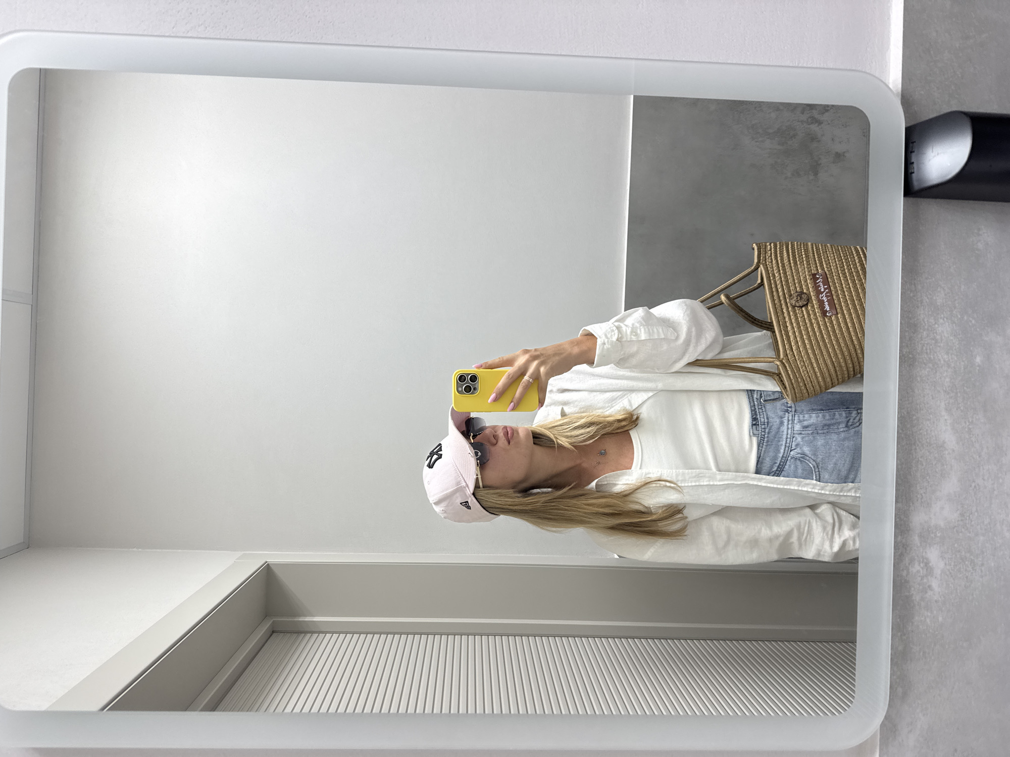 This is a mirror selfie taken in what appears to be a bathroom or dressing area. The subject, a woman with blonde hair, is wearing a white long-sleeved shirt, a white ribbed tank top, denim jeans, and a light pink baseball cap. She is also wearing sunglasses and has pink nail polish. She is holding a yellow smartphone to take the picture. A woven straw bag with a brown leather tag and a button closure is visible. The background of the reflection shows a white wall and a gray textured surface. The mirror itself has a rounded frame and appears to be backlit, creating a soft glow around the edges. A black object, possibly a hairdryer, is mounted on the wall to the right of the mirror. The lighting suggests it could be daytime, but the location and specific time are not evident. There is no discernible text visible in the image.