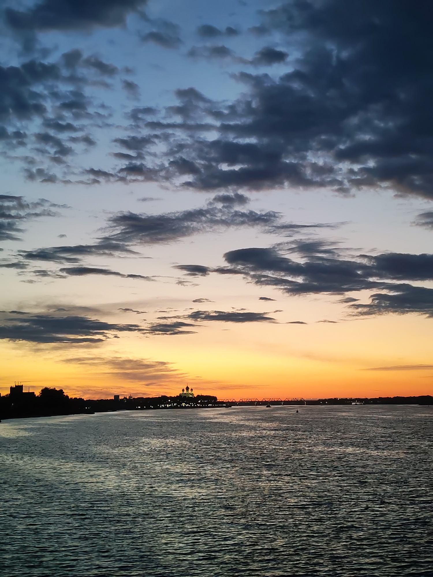 A wide shot captures a tranquil river scene at dusk in an unknown city. The sky is painted with hues of deep blue, gray, and vibrant orange, transitioning to a soft yellow near the horizon. Dark, scattered clouds drift across the sky, adding texture and depth. A silhouetted cityscape lines the distant shore, punctuated by architectural details that suggest buildings and possibly a church with domes. The surface of the river is dark and rippled, reflecting the colors of the sky. A bridge with several spans is visible in the distance, along with a few indistinct boats on the water. The overall impression is one of peacefulness and the calm beauty of twilight.