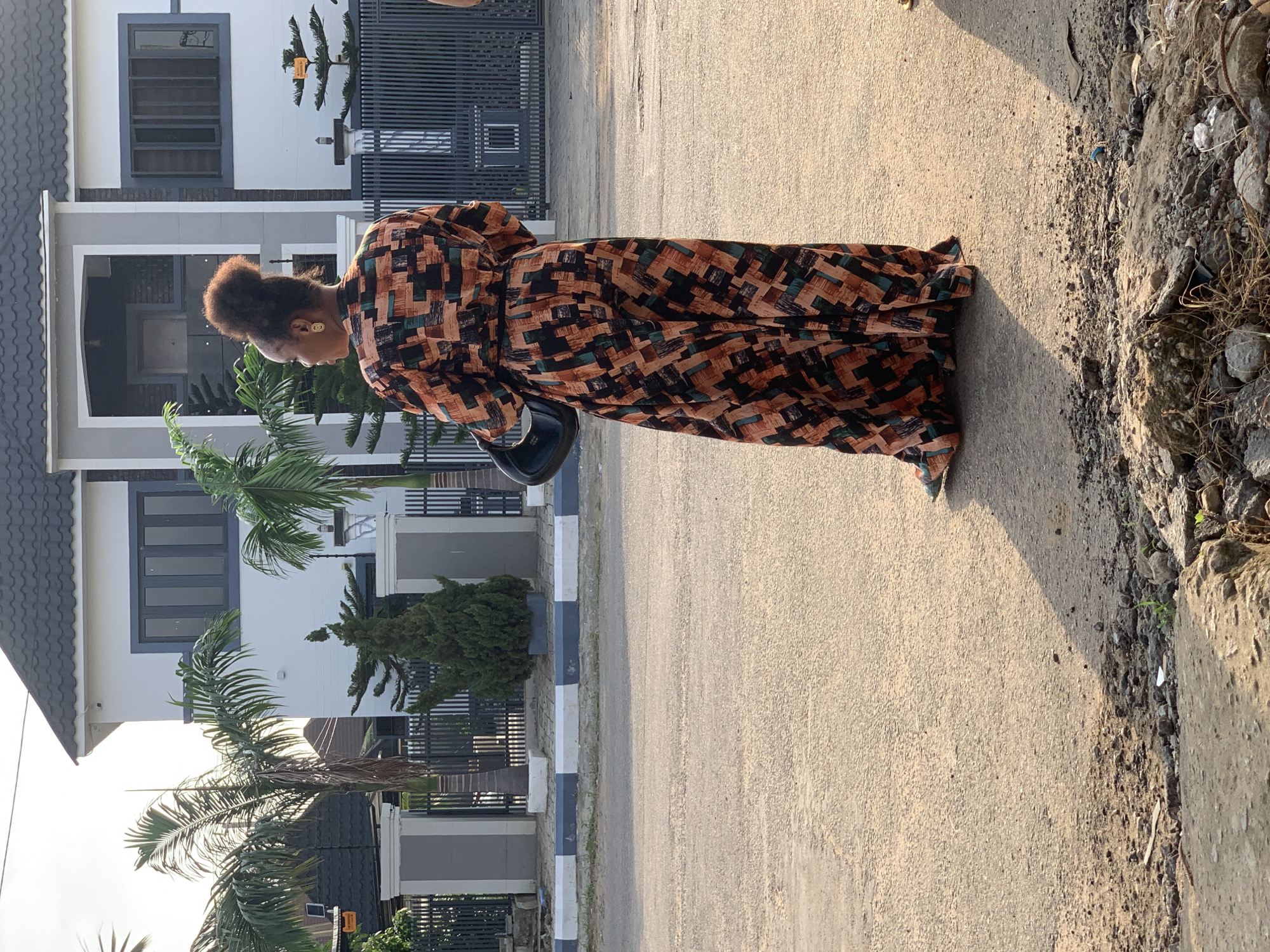 A woman is standing on an asphalt road in Abuja, Nigeria. She is facing away from the camera and is wearing a long, patterned dress with geometric shapes in orange, black, and teal. She has her hair pulled up in a bun and is wearing gold hoop earrings. A black bag is slung over her left shoulder. Behind her, a modern house with a white facade and dark grey roof is visible, along with palm trees and a metal fence. The road has a curb with alternating white and grey blocks. The sun is casting long shadows, indicating it is likely late afternoon or early morning. Debris and rocks are scattered on the side of the road in the foreground.