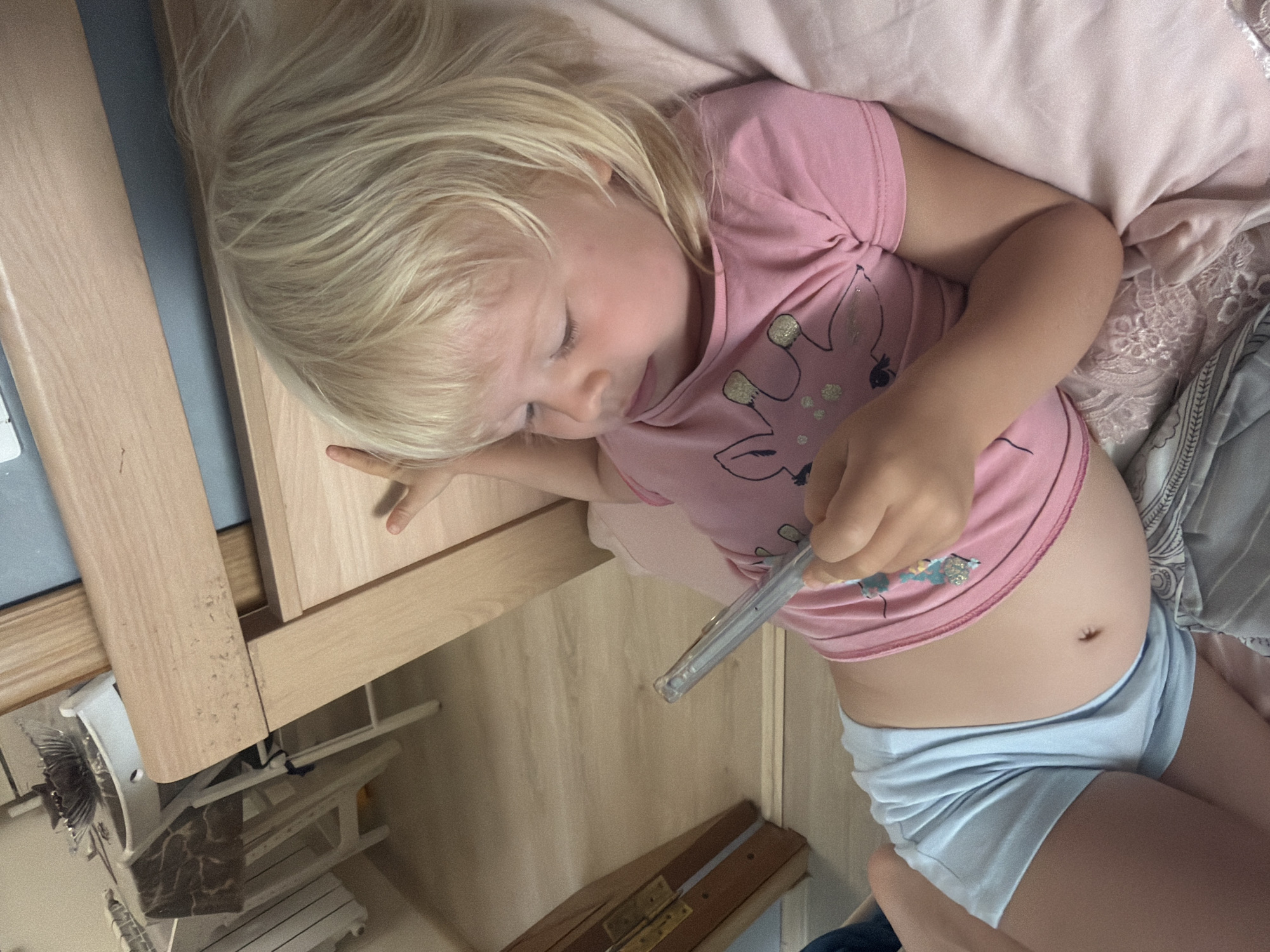 A young Caucasian child with blonde hair and bangs is lying on a bed, looking at a smartphone. The child is wearing a pink t-shirt with a graphic of a giraffe and light blue shorts. The background includes a wooden bed frame and a glimpse of a room with wooden flooring and what appears to be a radiator. The child is holding the phone in their right hand. The scene suggests a casual, domestic setting. There is no information to determine the specific location in the city of Sochi, Russia.