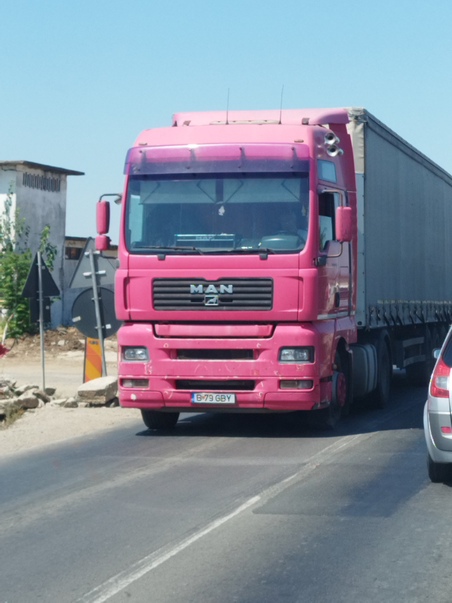 A bright pink MAN truck with a grey trailer is parked on the side of a road. The truck has a license plate that reads "B-79 GBY". A white car is visible on the right side of the frame. The background shows a building and some construction materials. The scene appears to be located in Clinceni, Romania, as indicated by the context. The road is paved and appears to be a public thoroughfare. The sky is clear and blue.