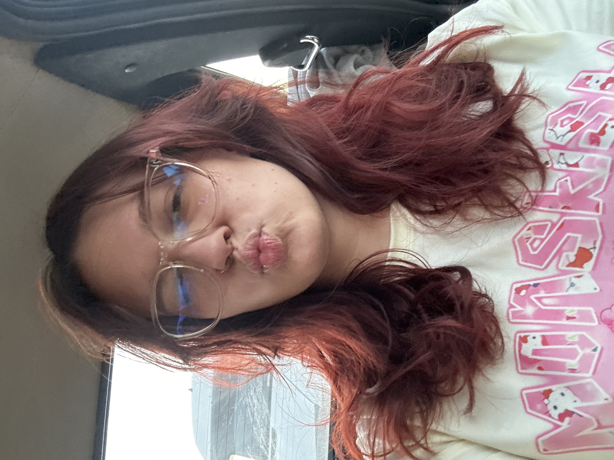 A young woman with long, wavy, dark red hair and glasses is in the driver's seat of a car, facing forward. She is making a puckering gesture with her lips and wearing a light-colored t-shirt with pink text and graphics. The background shows a window with a blurry view of what appears to be a street or another car, suggesting the vehicle is in motion. A car door handle and part of a seatbelt are visible on the right side of the frame. The scene is indoors, within a vehicle. There is no specific indication of the location within the city of Ленин, Kyrgyzstan.