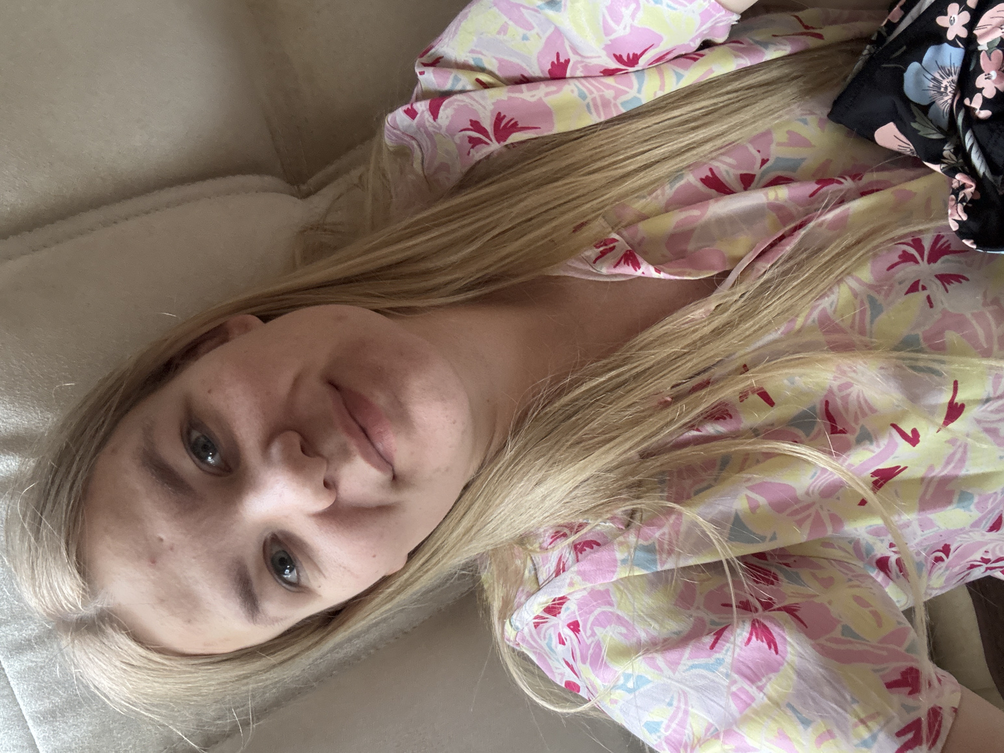 A fair-skinned young woman with blonde hair lies on a light-colored, possibly white, upholstered surface. She is wearing a light-colored floral print shirt with pink, yellow, and blue floral patterns. Another dark floral patterned garment is partially visible in the upper right corner. The woman has light blue eyes and a neutral expression. The photograph appears to be taken at a close-up, slightly tilted angle. The location within the city of Chaikovsky, Russia cannot be determined from the visual information.
