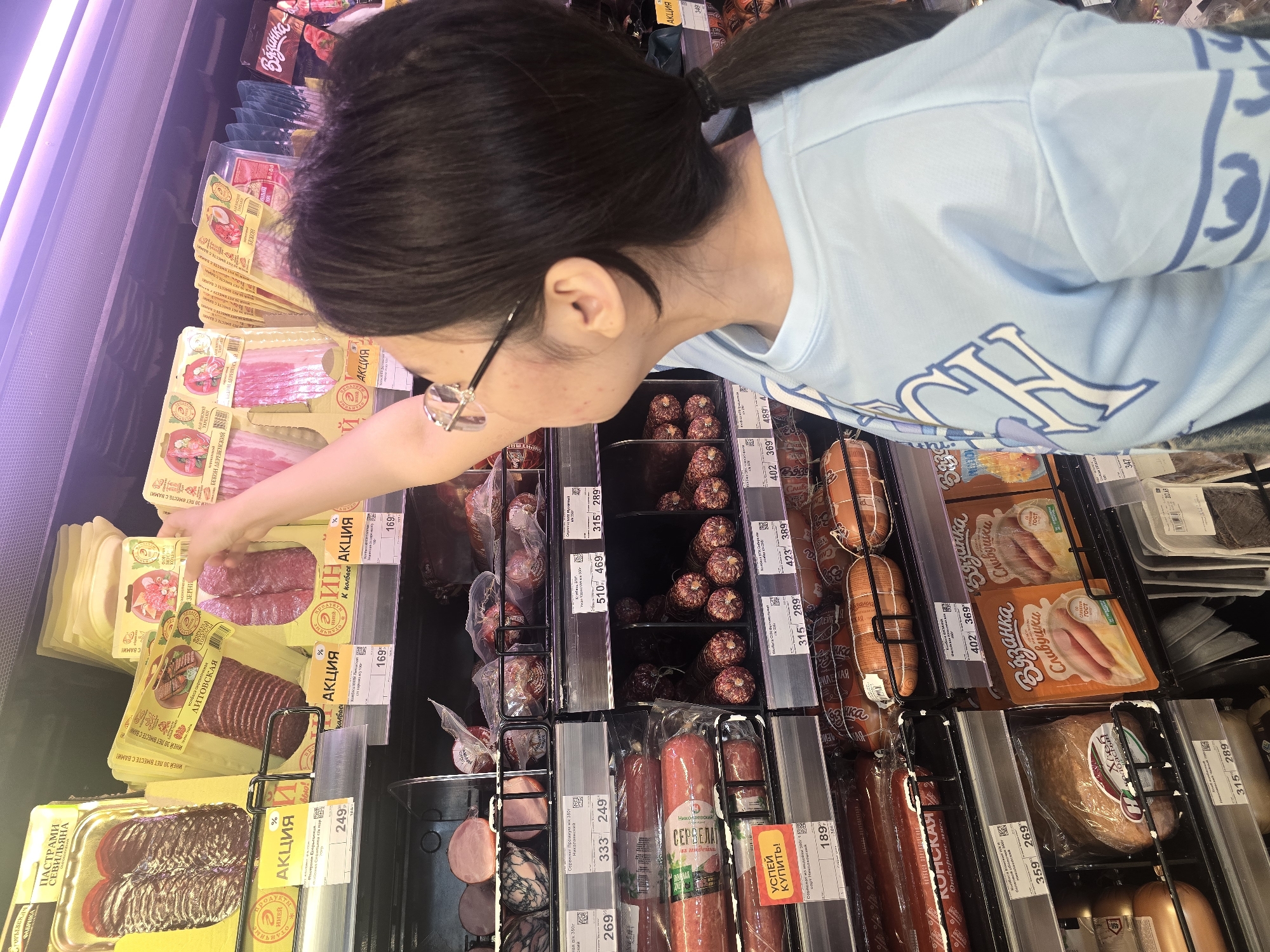 A young woman with dark hair pulled into a ponytail and wearing glasses and a light blue t-shirt is standing in a supermarket aisle, reaching for a package of sliced sausage from a shelf stocked with various meat products. The woman is looking up and to her right. The shelves are well-lit, displaying an assortment of packaged meats, including salami, ham, and bacon, with price tags and promotional labels visible. The environment appears to be a typical grocery store setting, with the shelves extending back into the distance. The overall impression is one of everyday shopping.