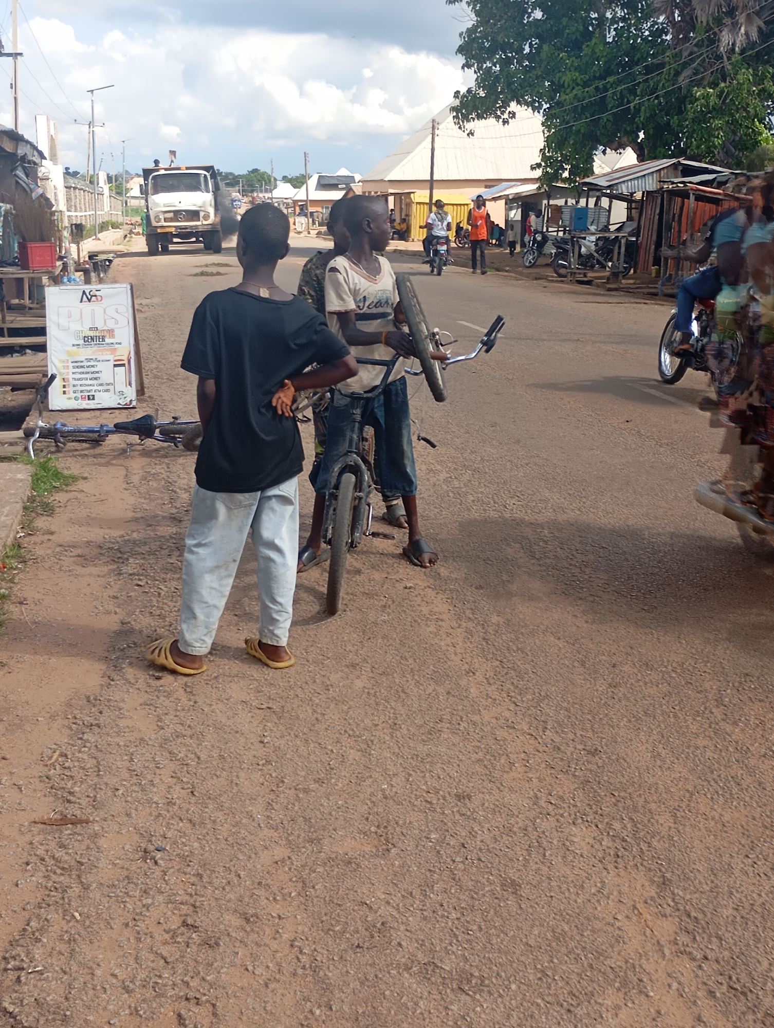 The image depicts a street scene in Shendam, Nigeria. In the foreground, three young males are visible. The leftmost individual, facing away from the camera, wears a black t-shirt and light-colored pants with yellow sandals. He has his left hand on his hip. To his right, another male, also facing away, wears a white t-shirt and dark pants. He is holding the handlebars of a bicycle. The third male, partially visible to the right, is wearing a light-colored t-shirt and dark pants, and appears to be standing near the second male. A bicycle lies on the ground to the left of the foreground figures. In the background, a white dump truck is driving down the road, emitting black exhaust fumes. Several roadside structures, including stalls with corrugated metal roofs, are visible on either side of the street. A signpost with information about a "POS Center" is situated to the left of the foreground figures. The ground is a dirt and gravel road. The sky is overcast with clouds.