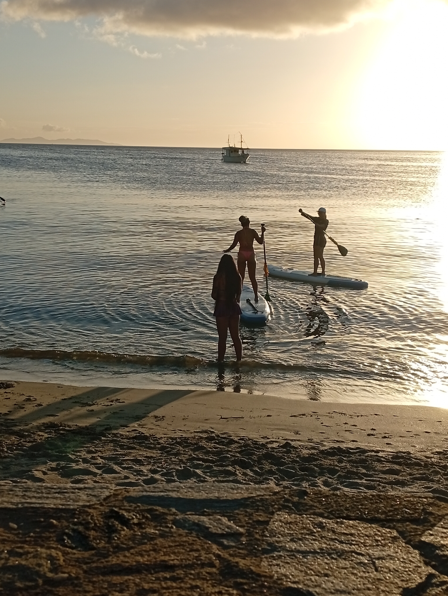 A wide shot captures a serene sunset scene at a beach. The sky is a soft gradient of pale yellow and orange, with a few wispy clouds. The sun, a bright disc, is partially obscured by a cloud. In the distance, a small boat with a single mast sails on the calm, rippling water. The horizon line is clearly defined by the sea and a distant island.
In the foreground, three figures are engaged in paddleboarding near the shore. The water is clear and reflects the warm light of the setting sun. One figure is standing closer to the shore, partially submerged in the shallow water, facing away from the camera. Two other figures are further out on their paddleboards. One is also facing away, while the other is looking towards the shore, with one arm raised as if gesturing. The sand in the immediate foreground is a light brown, with subtle footprints and shadows cast by unseen objects. The texture of the sand and some rocks are also visible. The overall atmosphere is peaceful and idyllic.