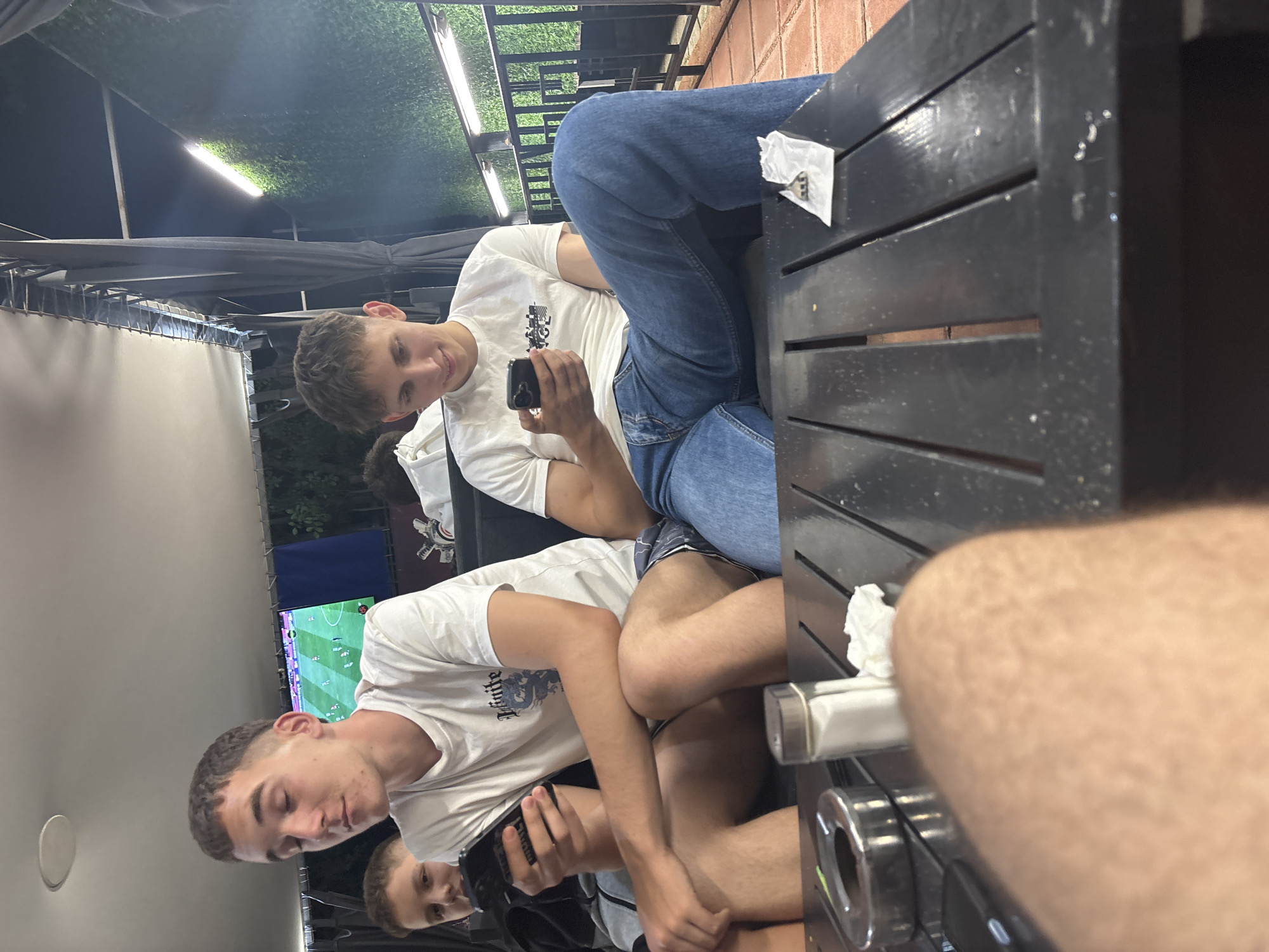 The image captures an evening scene at what appears to be an outdoor or semi-outdoor cafe or restaurant in Blagoevgrad, Bulgaria. Four young males are present, with the perspective taken from a low angle, revealing the photographer's blurred leg and arm in the immediate foreground.
In the middle ground, three males are seated on a bench or sofa. The central figure, a male in his late teens or early twenties with short brown hair, wears a white t-shirt featuring a dark graphic print, possibly "Infinite" with a dragon motif. He is engrossed in a black smartphone, holding it with both hands and looking down intently. He wears dark patterned shorts. To his left, partially visible behind his shoulder, is another male with lighter hair, also looking downwards, suggesting engagement with a personal device. This individual appears younger, likely in his mid-teens.
To the right, a third male, also in his late teens or early twenties, with short, curly light brown hair, smiles faintly as he looks down at a black smartphone held in his left hand. He wears a white t-shirt with a dark, indistinct graphic print on the chest and blue jeans.
A dark, slatted table occupies the lower third of the frame. On its left side, a metallic ashtray with a bright green rim is visible. Next to it, a clear salt shaker filled with white crystals stands upright. Further right on the table, a small white paper packet, possibly containing sugar or a wet wipe, has a small metallic utensil (resembling a miniature fork or spoon) resting on it.
The background shows artificial lighting against a dark sky. To the left, a flat-screen television mounted on a wall displays a football (soccer) match in progress. To the right, a dark green artificial hedge wall is illuminated by two prominent horizontal strip lights. Above, a white canopy structure supported by dark metal beams and draped with dark fabric curtains indicates a covered outdoor area. The overall lighting suggests it is nighttime or late evening.