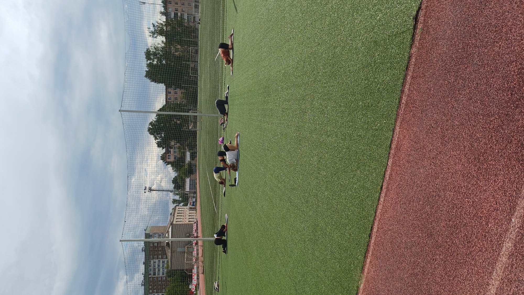 A group of five individuals are engaged in yoga poses on a green artificial turf field, likely a sports stadium, under a cloudy sky. Buildings and trees are visible in the background. One person is in a quadruped position facing left, another is in a similar position with their head down, a third is on their hands and knees with their head down, a fourth is on their knees facing away from the camera, and the fifth is in a quadruped position facing right. The image is captured from a slightly elevated perspective. The location is identified as Izhevsk, Russia.