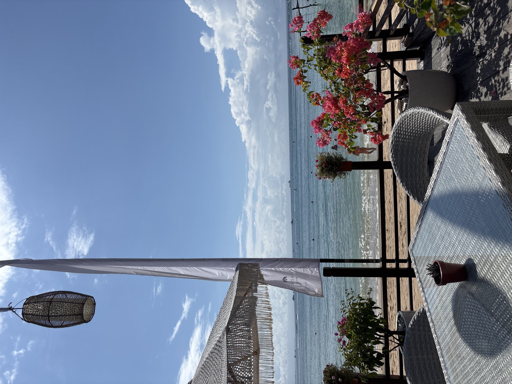 The scene is an outdoor setting in Kuta Selatan, Indonesia, likely a beachfront restaurant or lounge, as suggested by the view of the ocean, sandy beach, and the presence of a covered seating area.
The sky is predominantly blue with some scattered white clouds. A long, white canopy is seen stretching across the frame. Beneath the canopy, a decorative lamp hangs.
The right side of the image features a wooden deck with tables and chairs. The tables are white wicker with glass tops. There are potted plants with pink flowers on the deck.
The ocean is visible in the background, with small vessels visible in the distance.