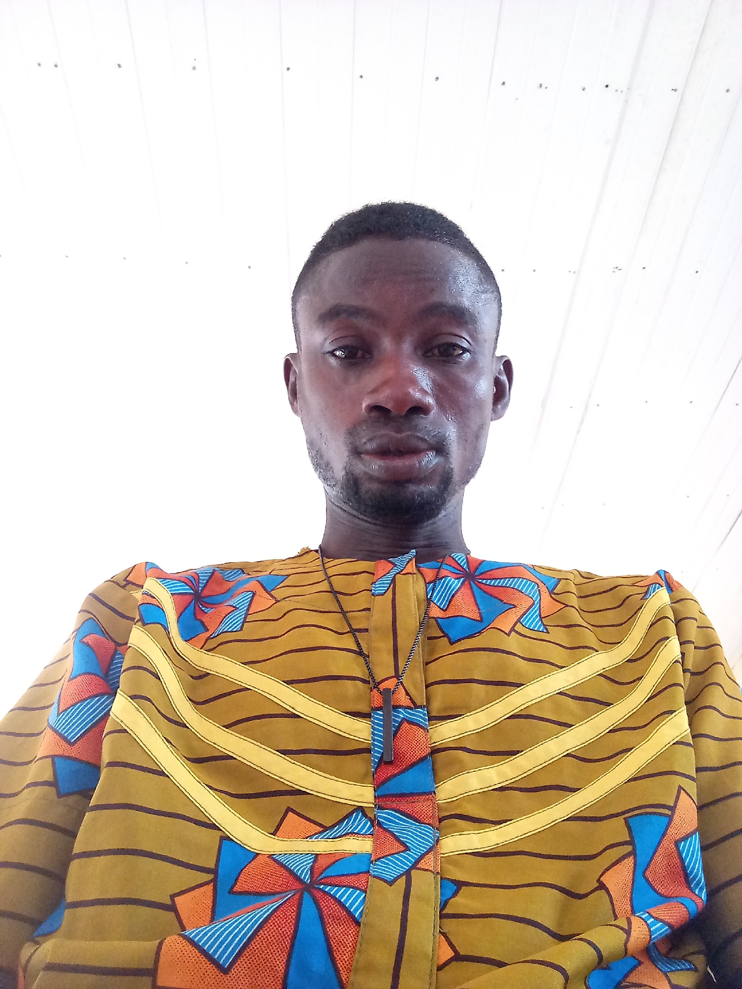 A man stands in front of a white, paneled ceiling, wearing a patterned shirt. The shirt has a base color of olive green, with horizontal dark brown lines and yellow stripes. There are large, flower-like patterns with orange, red, and blue geometric designs. The man is wearing a necklace with a dark rectangular pendant. The location is implied to be Zango-ndaji, Nigeria.