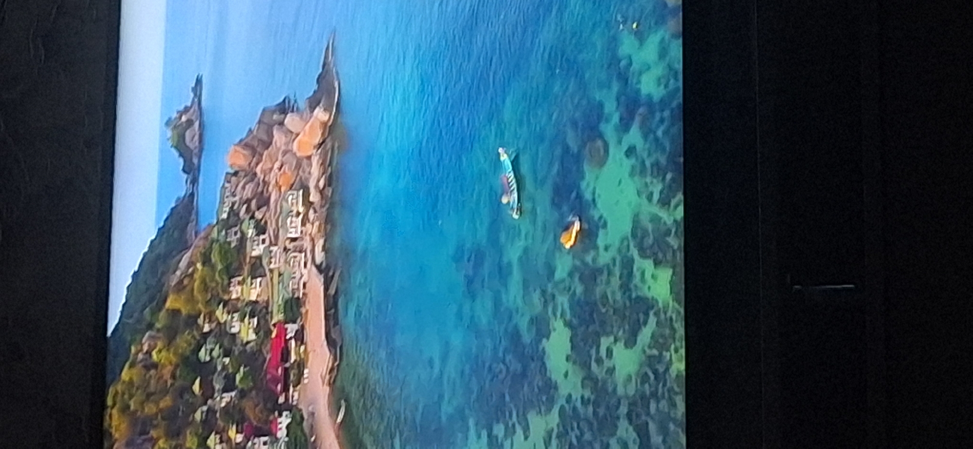 This image depicts a picturesque coastal scene with a cluster of white and red-roofed buildings nestled on a hillside overlooking a clear, turquoise ocean. The buildings appear to be villas or small resorts, with green rooftops. The coastline is rocky, with lush vegetation covering the hills. In the water, there are two small boats, one longer and blue-and-white striped, and another smaller, round, orange object that could be a buoy or inflatable. The water is shallow enough in some areas to see the coral or rock formations beneath the surface, creating a mosaic of green and blue hues. In the distance, a small rocky island with some vegetation rises from the sea. The overall impression is one of a tranquil tropical or Mediterranean-like destination. There is no indication of the city of Bila Tserkva, Ukraine, in this image.