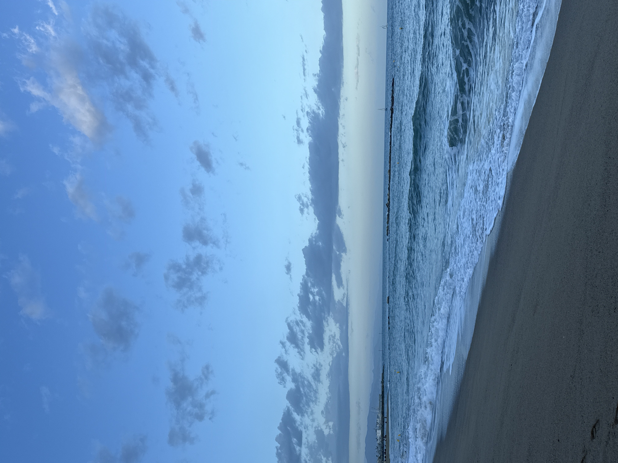 This is a daytime view of a beach in Barcelona, Spain, with the sea on the right side of the frame, and the sky on the left. The sea is choppy with waves. A breakwater extends into the sea in the middle ground. The beach is composed of dark sand. The sky contains scattered clouds. There are distant buildings visible on the horizon in the middle ground. Footprints are visible on the sand.