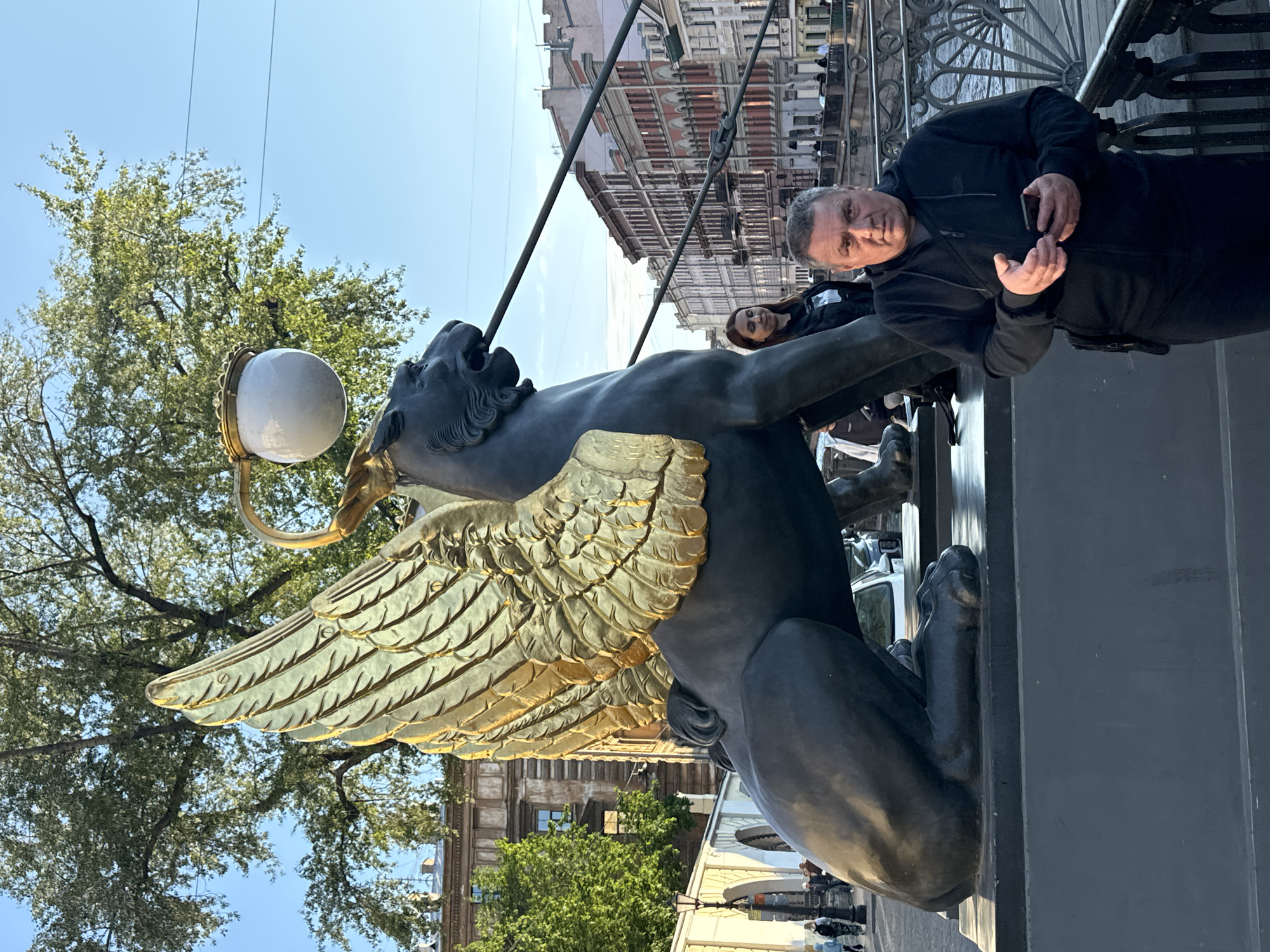In Saint Petersburg, Russia, a man stands in the foreground of the image, holding a smartphone. He is positioned in front of a large, dark sculpture of a griffin with golden wings, which appears to be a lamp post, with its beak open and a lamplight above. Behind the griffin, a woman in a black jacket is visible, and further in the background, a row of classical buildings lines a canal, with a bridge railing and the canal's water also present. The scene is set against a backdrop of trees and a clear blue sky.