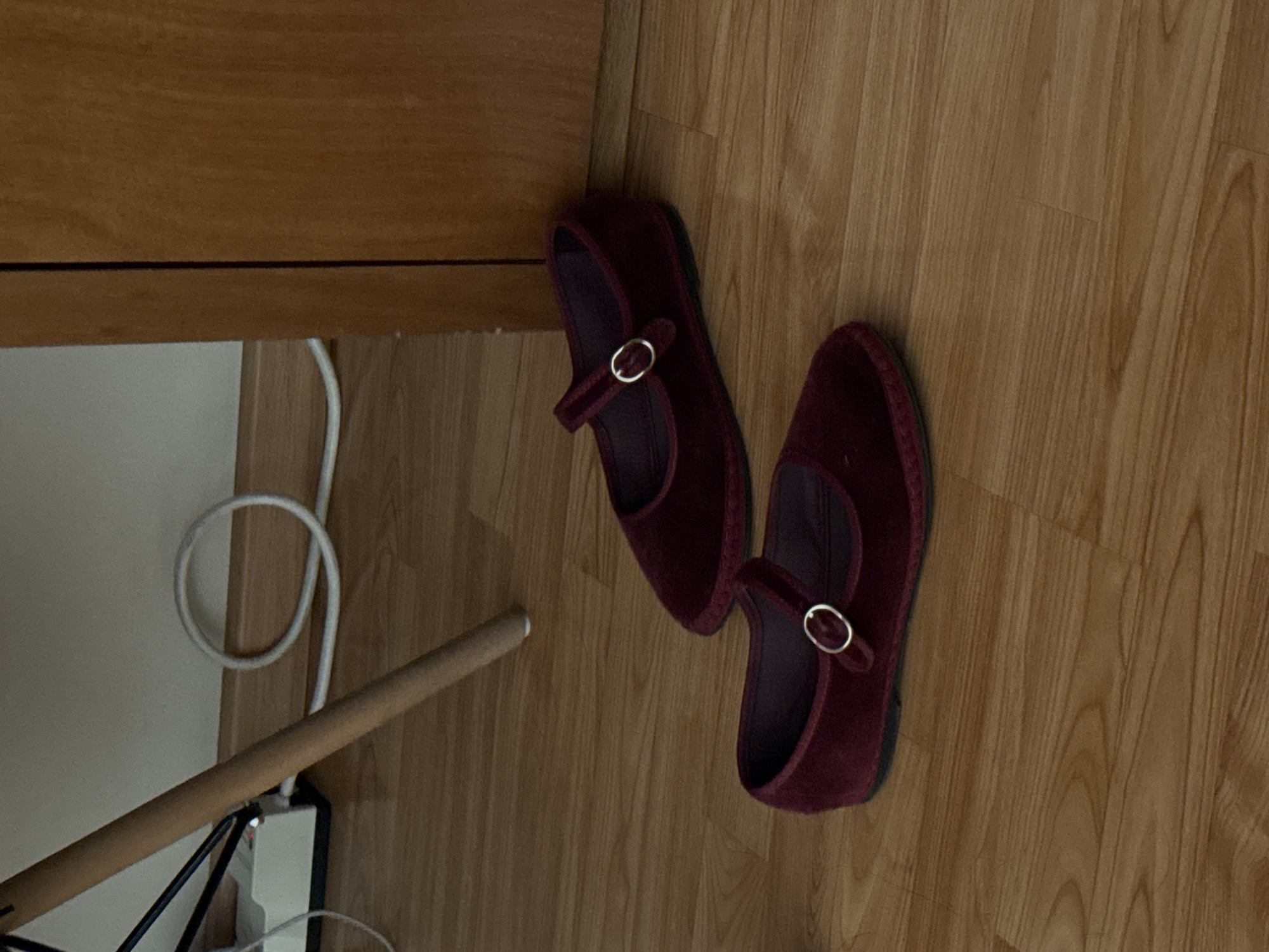 This image, captured in San Bernardino Tlaxcalancingo, Mexico, presents an indoor scene with a focus on footwear and flooring. The central subjects are a pair of maroon or burgundy Mary Jane-style shoes, likely made of velvet or suede, each featuring a single strap across the instep secured by a small, round, gold-colored buckle. The shoes are placed on a light-toned wooden floor, which displays prominent grain patterns.
In the upper left portion of the frame, a segment of light brown wooden furniture, possibly a desk or cabinet, is visible. Below this furniture, a white wall runs vertically. A coiled white electrical cable lies on the floor near the wall. Towards the bottom left, a smooth, light-brown wooden pole or chair leg extends diagonally. Partially obscured behind this pole is a white power strip, with various dark cables plugged into it. The overall setting is a casual, domestic environment. No people are visible, and no specific activity is taking place beyond the placement of the shoes. The lighting suggests an indoor setting, with no indications of the time of day or external weather. There is no visible text in the image.