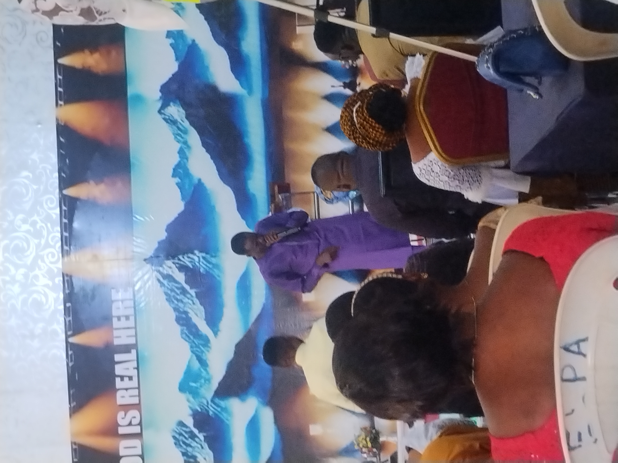 A partially obscured image captures a gathering of individuals inside a building in Kabong, Nigeria. A man in a purple suit stands on what appears to be a stage or platform, holding an object resembling a microphone or small device. Behind him, a large banner with text "GOD IS REAL HERE" is displayed with a backdrop of a mountain scene. Several people sit in chairs facing the man on stage, with partial views of their clothing (red, white, yellow). The environment appears to be an interior space, likely a church or event hall based on the context provided by the banner.