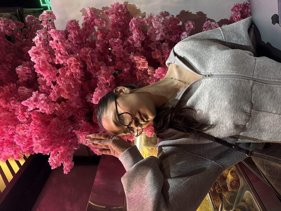 A female individual is depicted indoors in Yerevan, Armenia. She is observed from the chest up, positioned slightly left of center, her head tilted back and to the right with a faint smile and partially closed eyes. She has dark hair pulled back and wears dark-rimmed eyeglasses, a grey hooded sweatshirt with a front zipper, and a thin gold-colored necklace. Her right hand is raised, palm open and fingers extended, resting against an opaque vertical surface. The background on the right is dominated by a dense, vertical arrangement of artificial pink flowers. To her left, a commercial display case contains various pastries and sweets, above which a reflective metallic surface and a ceiling with dark and yellow striped elements are visible. The scene is illuminated by artificial light.