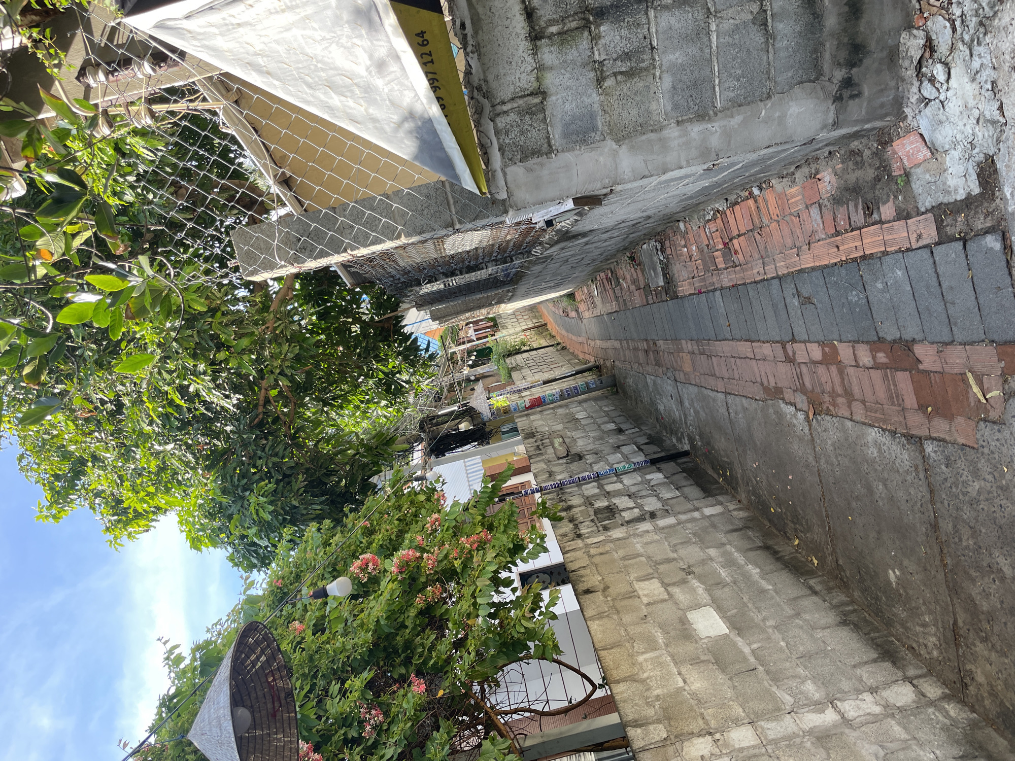 The image depicts a narrow alleyway in Phu Thuy Ward, Vietnam. The alley is lined with aged concrete block walls on both sides. The ground is a mix of concrete and red brick paving. Lush green trees with pink flowers overhang the alley from the left, with a conical woven hat lamp and a white light bulb hanging from wires above. On the right, a chain-link fence runs along the top of a concrete wall, behind which a building with a yellow facade is visible. A white banner with black text and a yellow stripe, featuring the numbers "997 12 64", is attached to the fence. Electrical poles with wires and various posters are also present. The sky above is blue with some white clouds. The scene suggests a residential or commercial area with elements of tropical vegetation.